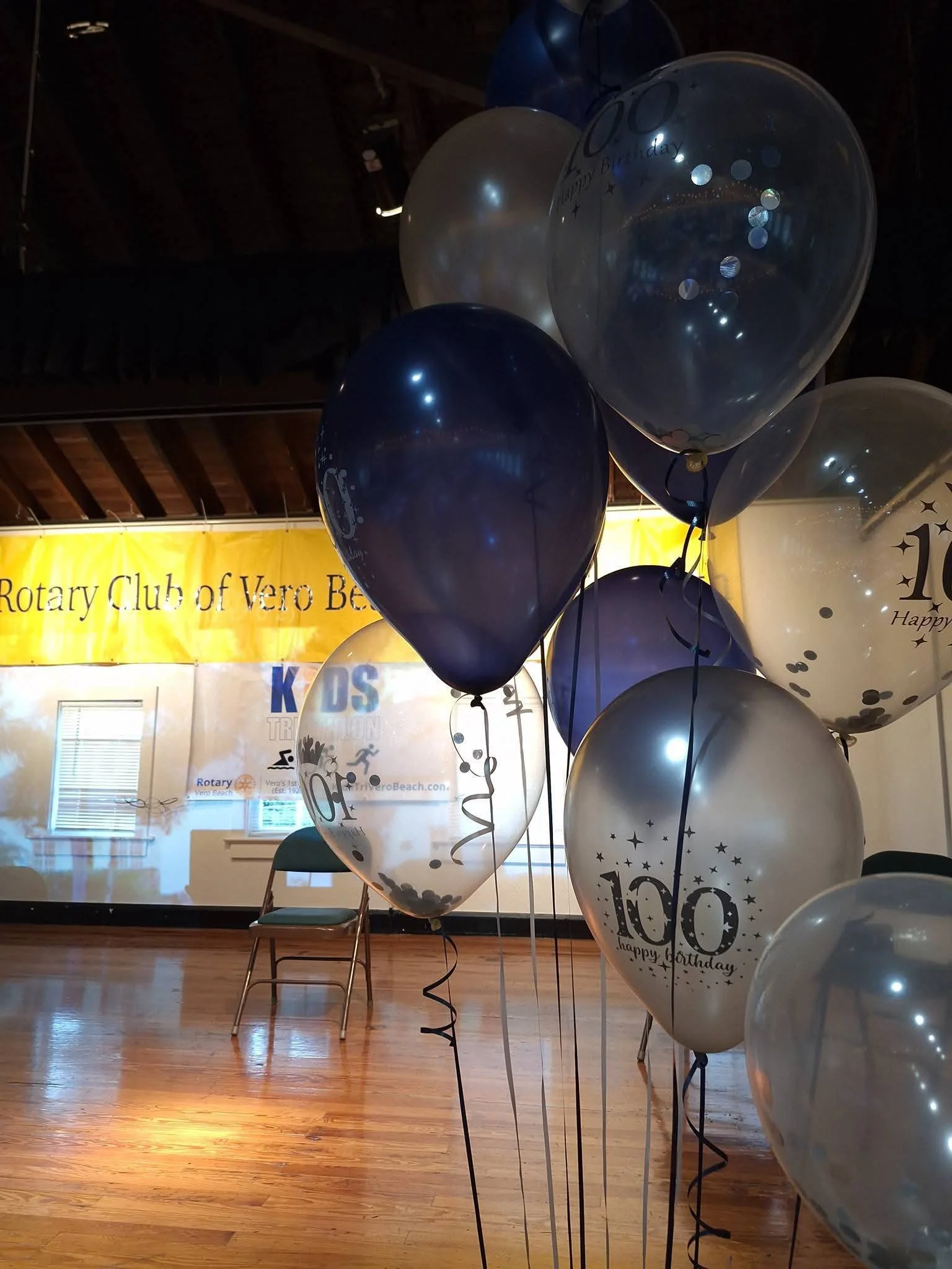 Balloon decorations with blue, white, and silver balloons, some with printed birthday messages, set over a wooden floor at the Rotary Club of Vero Beach.