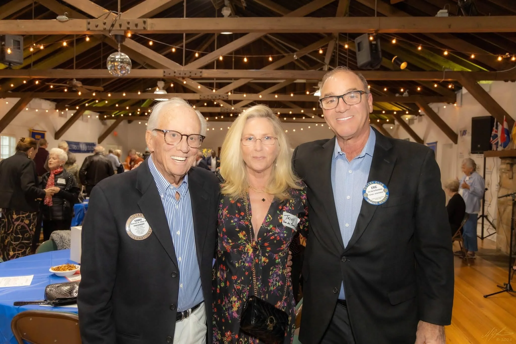 Three people smiling at a social event in a large hall with string lights and wooden beams. The man on the left is wearing glasses, a badge that says he is the president of Rotary International, and a dark blazer. The woman in the middle has blonde hair, glasses, and is wearing a colorful floral blouse with a name tag. The man on the right is wearing glasses, a dark blazer with a Rotary badge, and a light blue shirt.