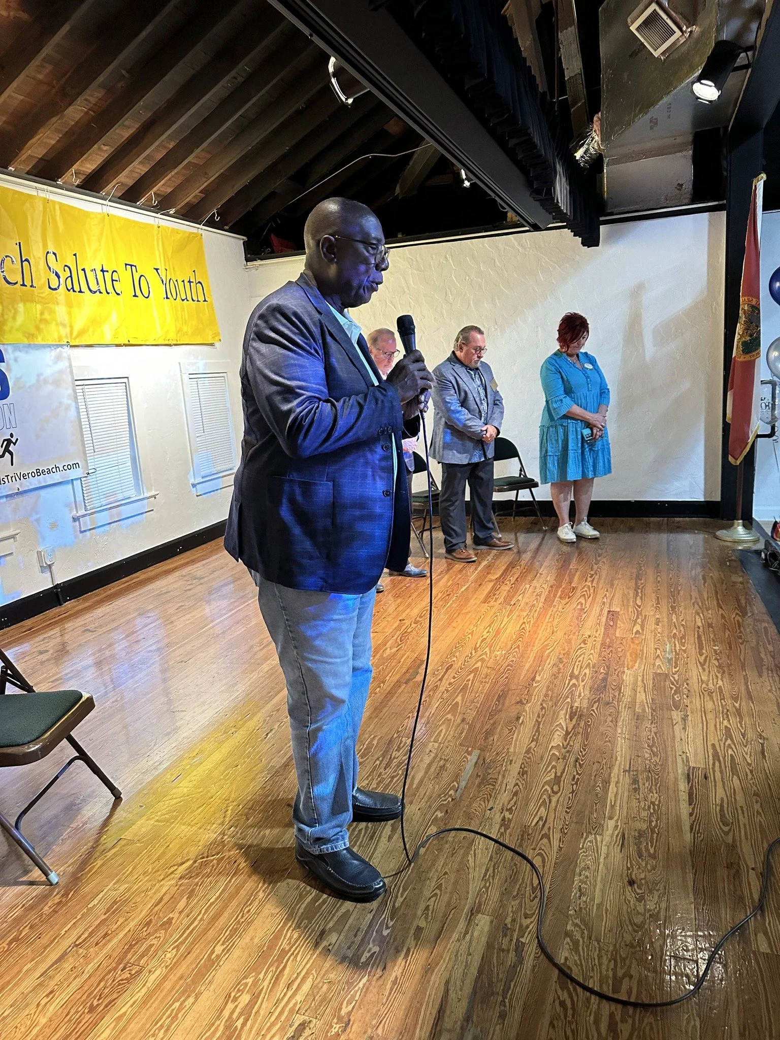 A man in a gray checkered blazer and glasses stands holding a microphone, reading or speaking at an event. In the background, three other people stand with their heads bowed. There is a yellow banner with partially visible text that reads '...ch Salute To Youth' on the wall behind them. The setting is an indoor venue with wood flooring and a ceiling with exposed wooden beams.