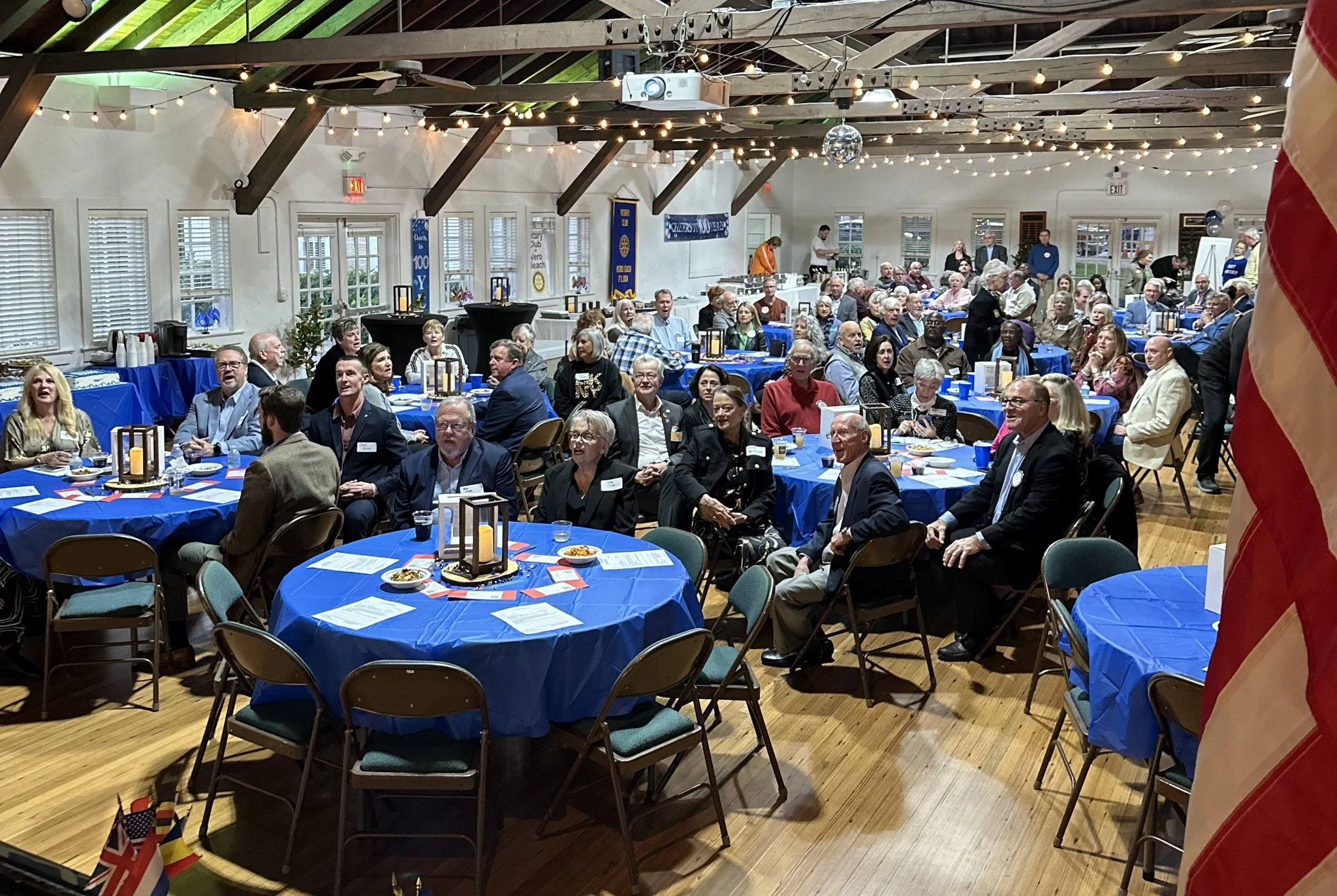 Indoor event with people seated at round tables covered with blue tablecloths, decorated with lanterns and American flags, under string lights and wooden beams.