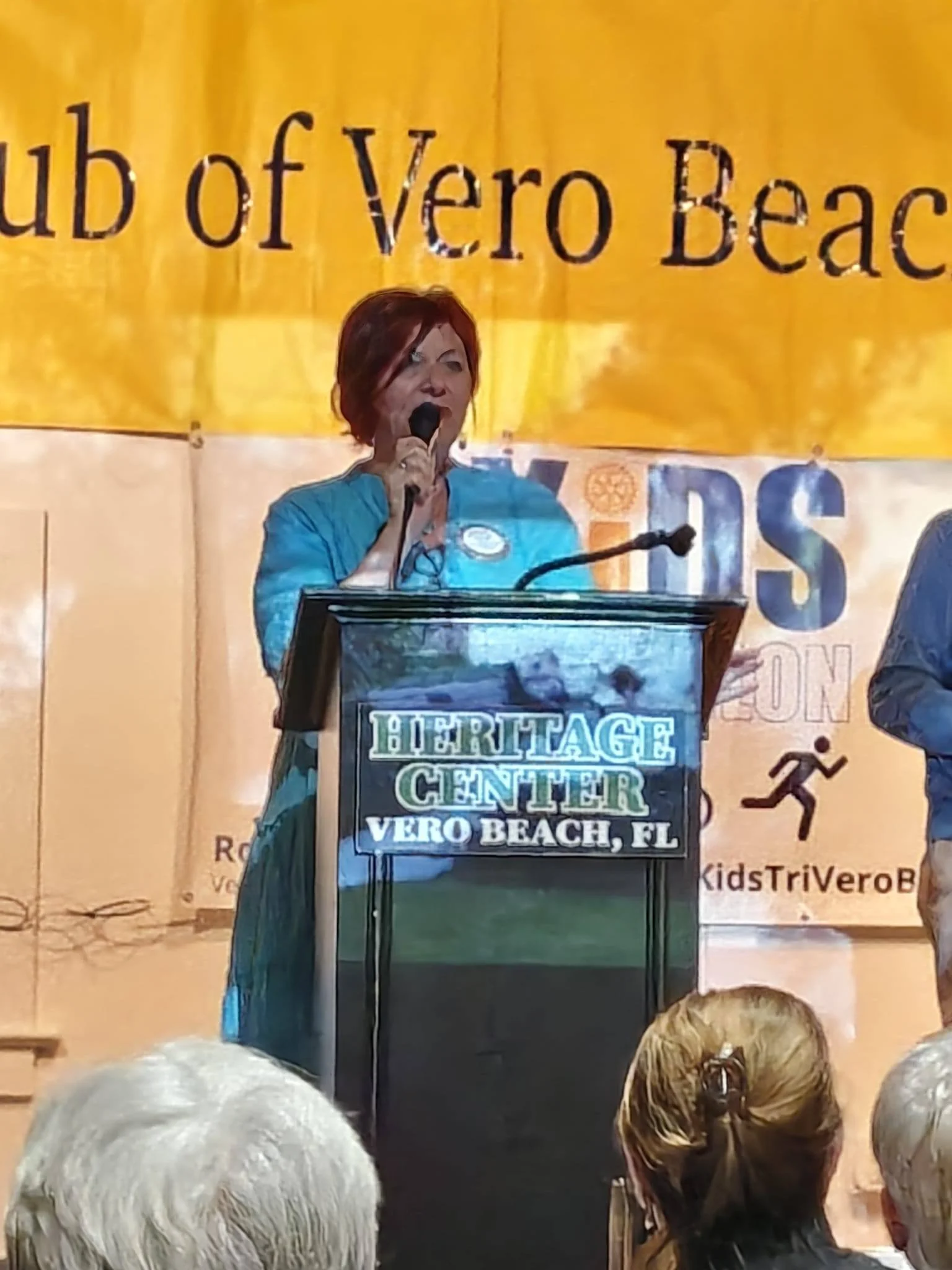 Woman with short red hair standing at a podium, speaking into a microphone during an event at the Heritage Center in Vero Beach, FL, with an audience of mostly older individuals visible in the foreground.