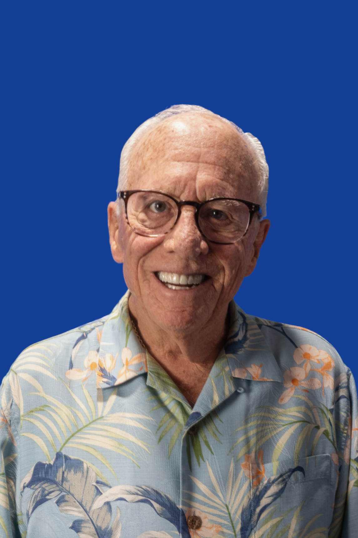 An elderly man with glasses, white hair, and a suit holding a puzzle box, smiling at a social gathering with warm lighting and a blue banner in the background.