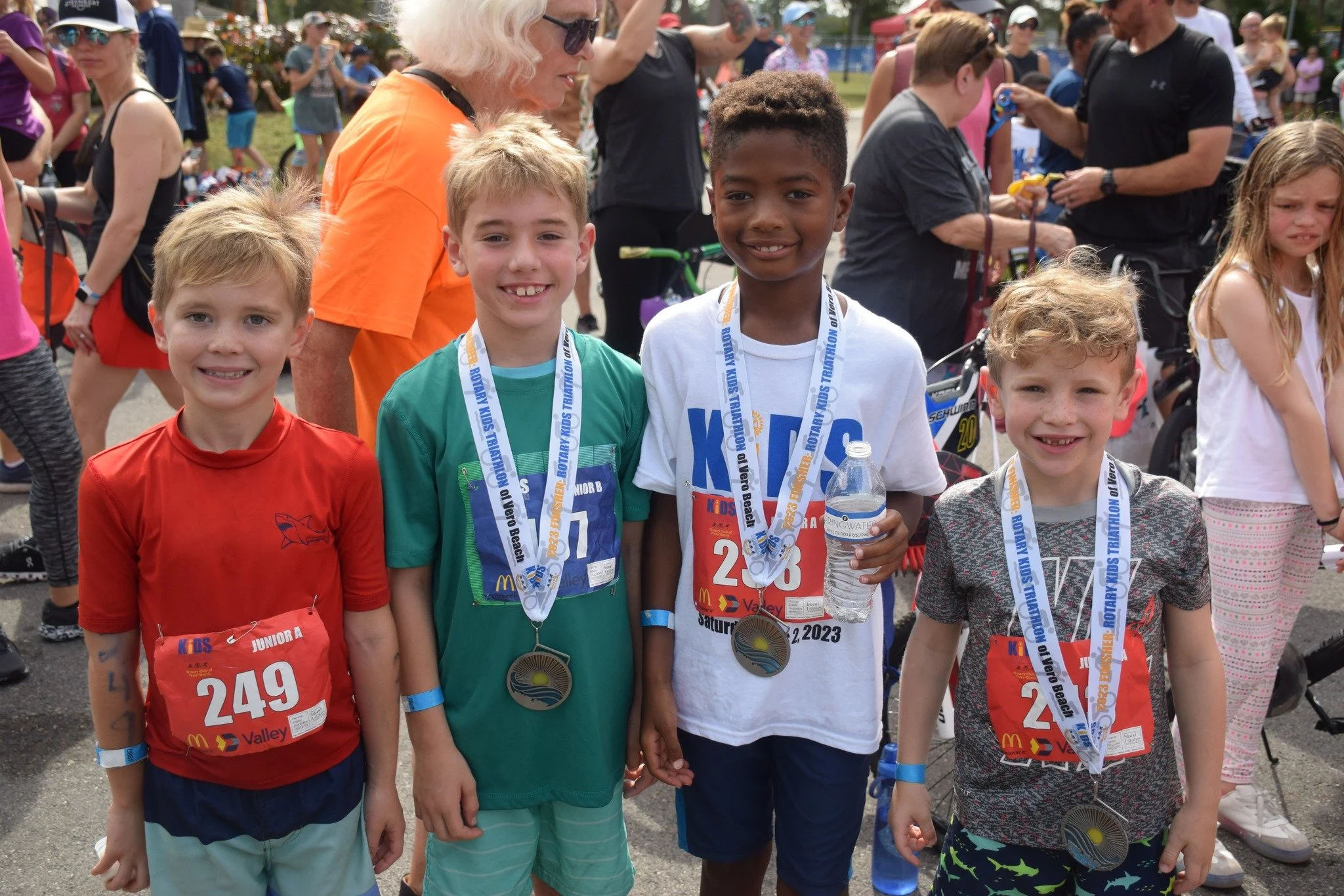Four young boys with medals and race bibs, smiling at a race event, surrounded by other participants and spectators.