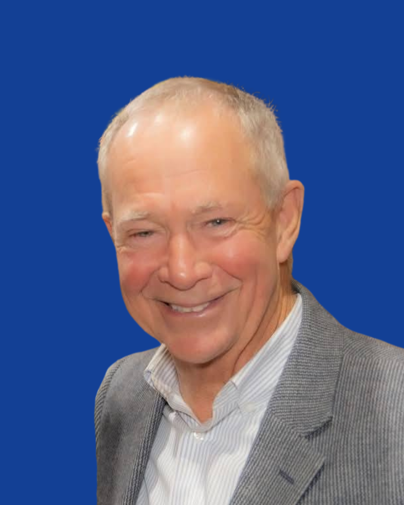 A smiling older man wearing a gray blazer and white dress shirt at a gathering indoors with warm lighting and string lights.