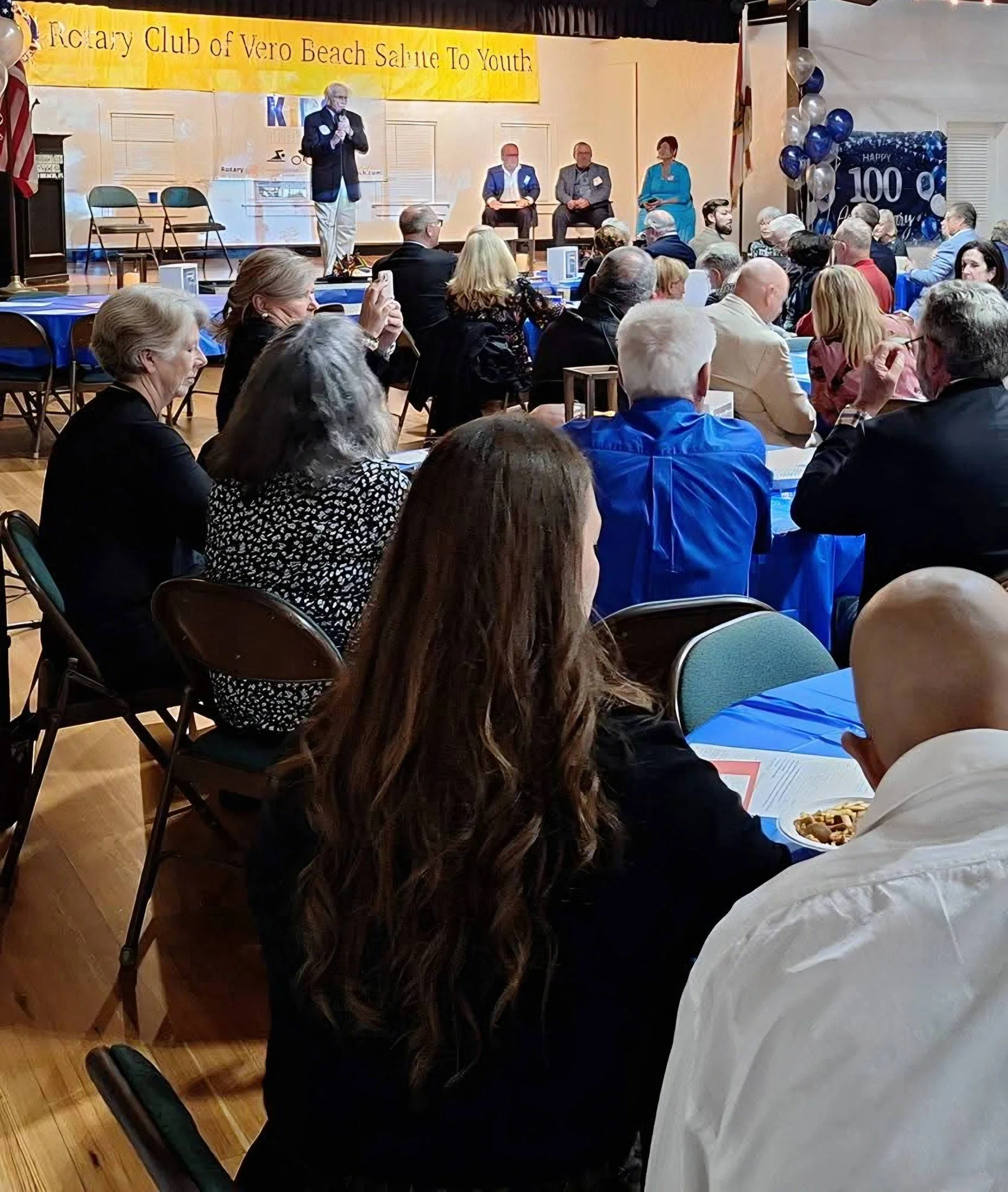 Indoor event space with people seated at tables covered in blue tablecloths, some eating, observing a stage with a speaker, American flag, and balloons. A yellow banner reads 'Realtor Club of Vero Beach Salute To Youth'. Decorations include balloons and a sign celebrating a 100th anniversary.