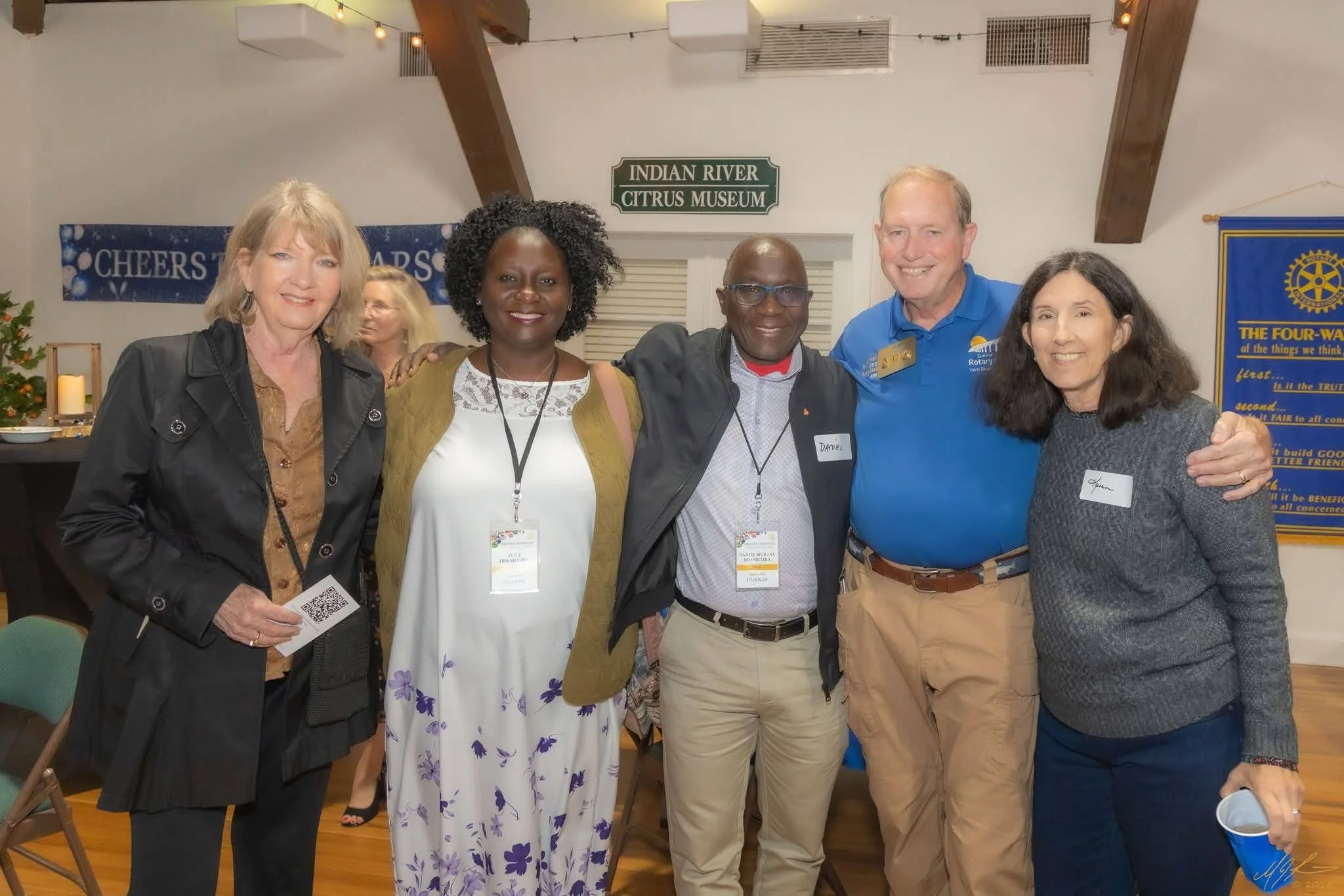 Group of five diverse adults smiling and standing together at an indoor event, with signs and banners in the background, including one that reads 'Indian River Citrus Museum'.