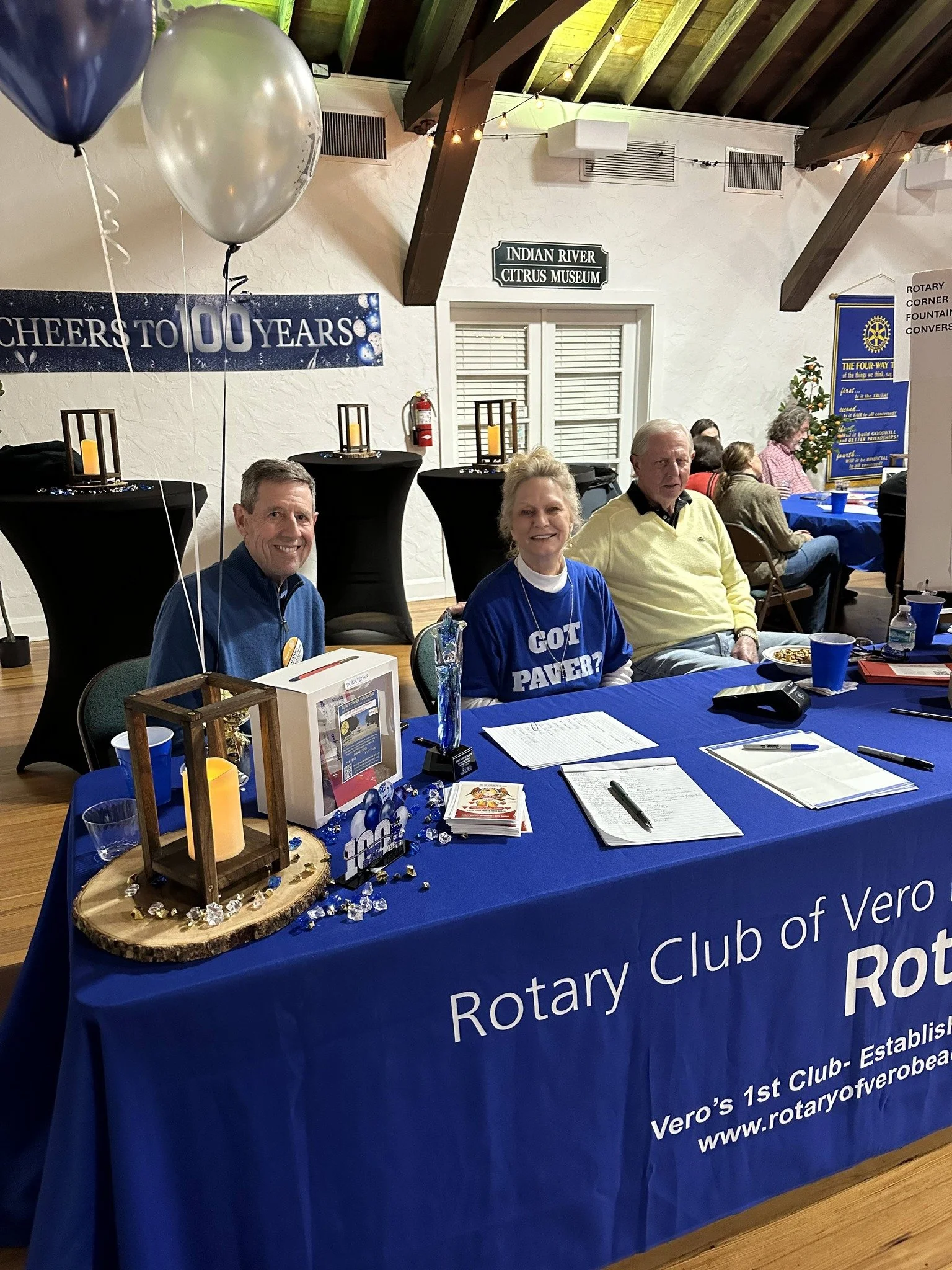A Rotary Club of Verobeach table with a blue tablecloth, displaying various flyers, a small wooden lantern, and decorative items. Behind, three people are seated at the table, smiling. The background has balloons, a banner celebrating 100 years, and other Rotary Club banners, with a wood-beamed ceiling and white walls.