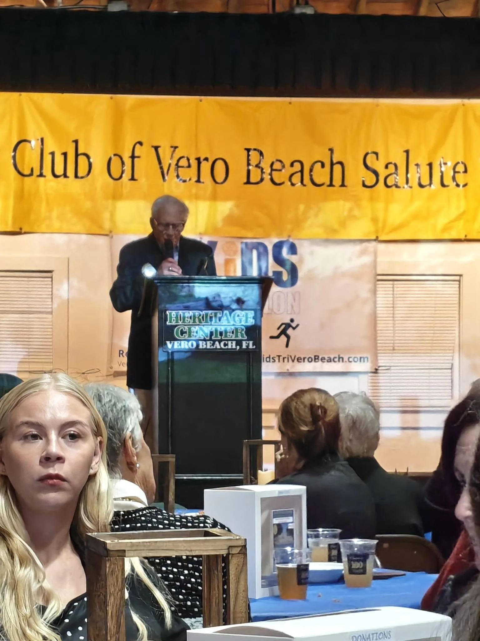 A man giving a speech at a podium in front of a yellow banner that reads 'Club of Vero Beach Salute' at the Heritage Center in Vero Beach, Florida. Several people are seated, listening, in the foreground.