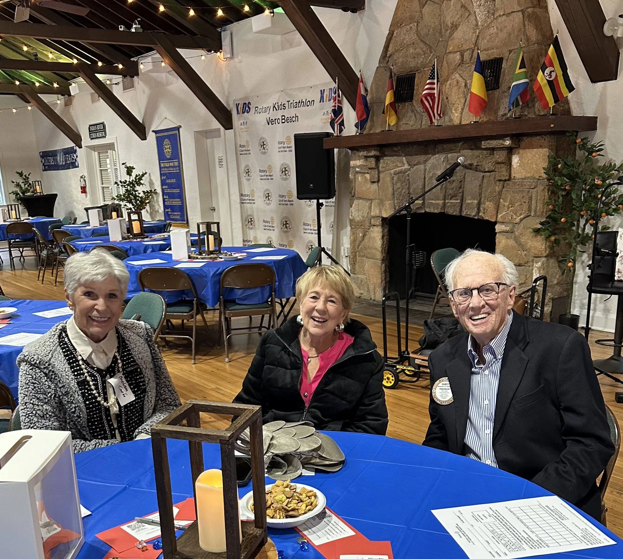 Group of smiling older adults seated at decorated tables during an indoor event, with a stage and flags in the background.