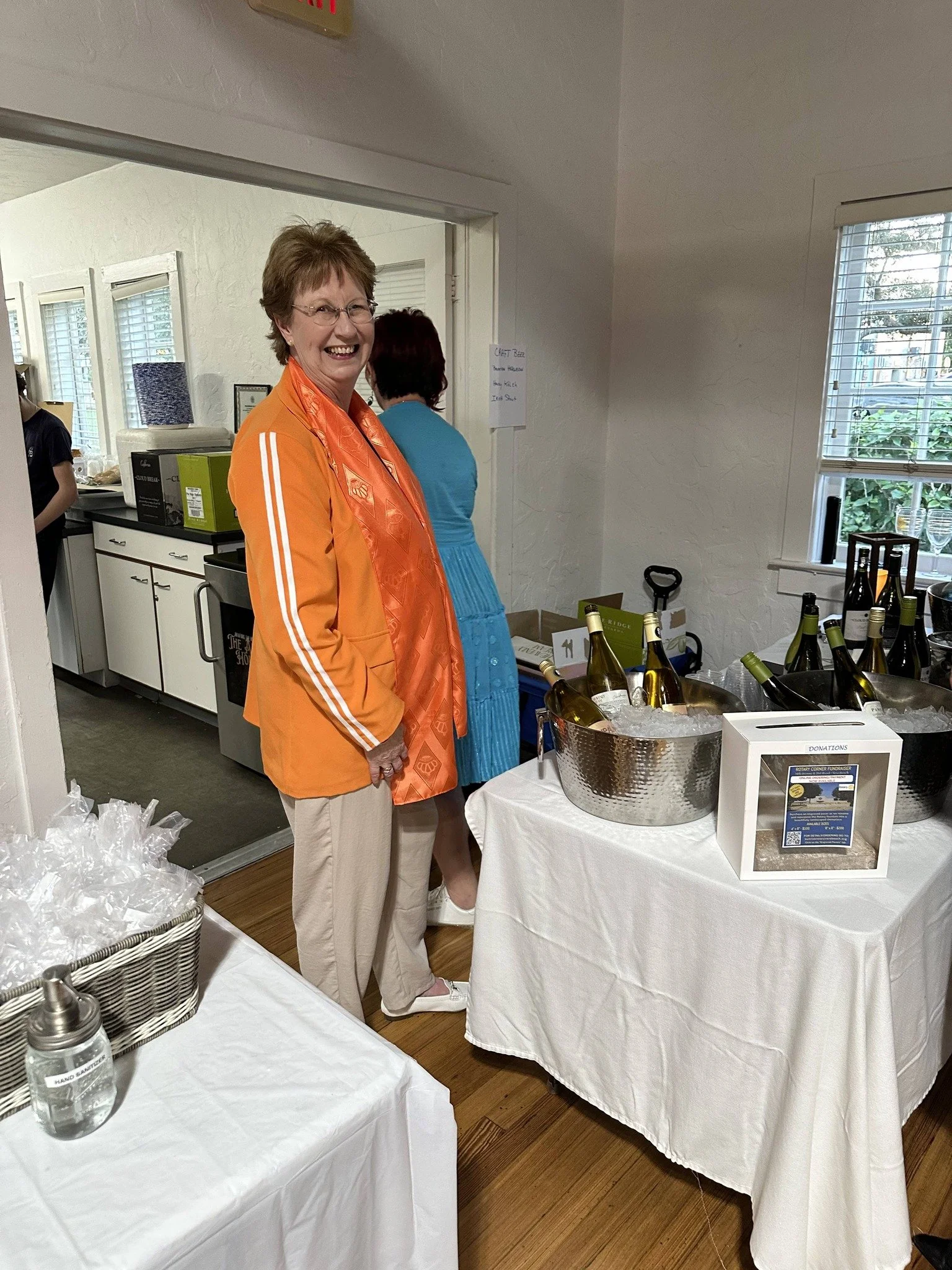 A smiling woman in an orange jacket and beige pants stands beside a table with wine bottles, ice, and a donation box, in an indoor setting with white walls and windows.