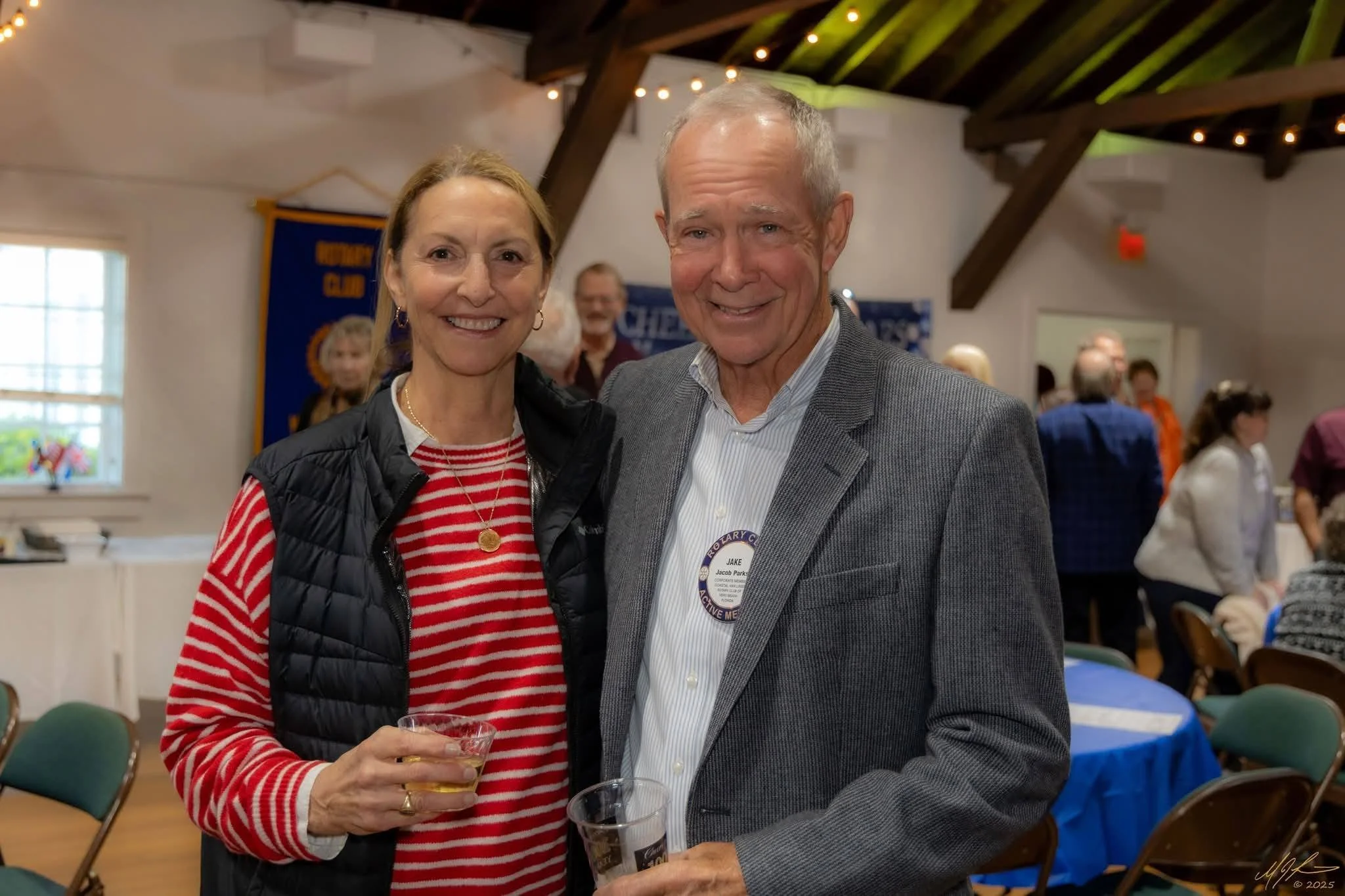 A woman and a man smiling and standing close together at an indoor gathering. The woman is wearing a red and white striped shirt and a black vest, holding a drink, and the man is dressed in a gray suit jacket and holding a drink. The background shows other people, a blue banner, and decorative string lights in a room with wooden beams.