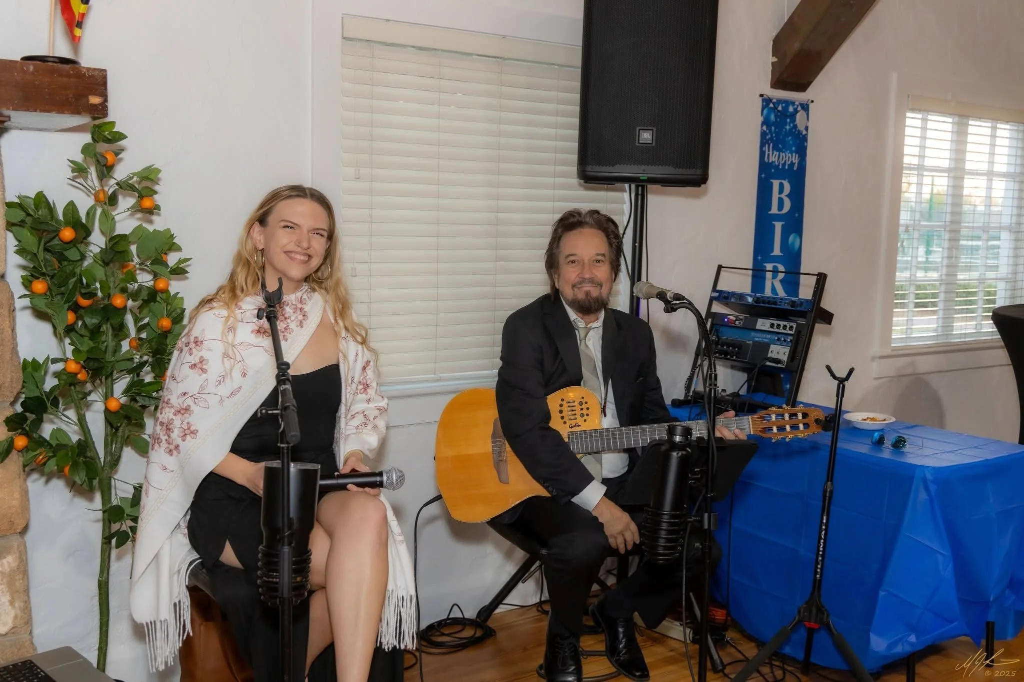 A woman sitting on a chair holding a microphone, smiling, next to a man playing an acoustic guitar, both in a room decorated with a small orange tree, a blue banner, and various music equipment on a blue table.