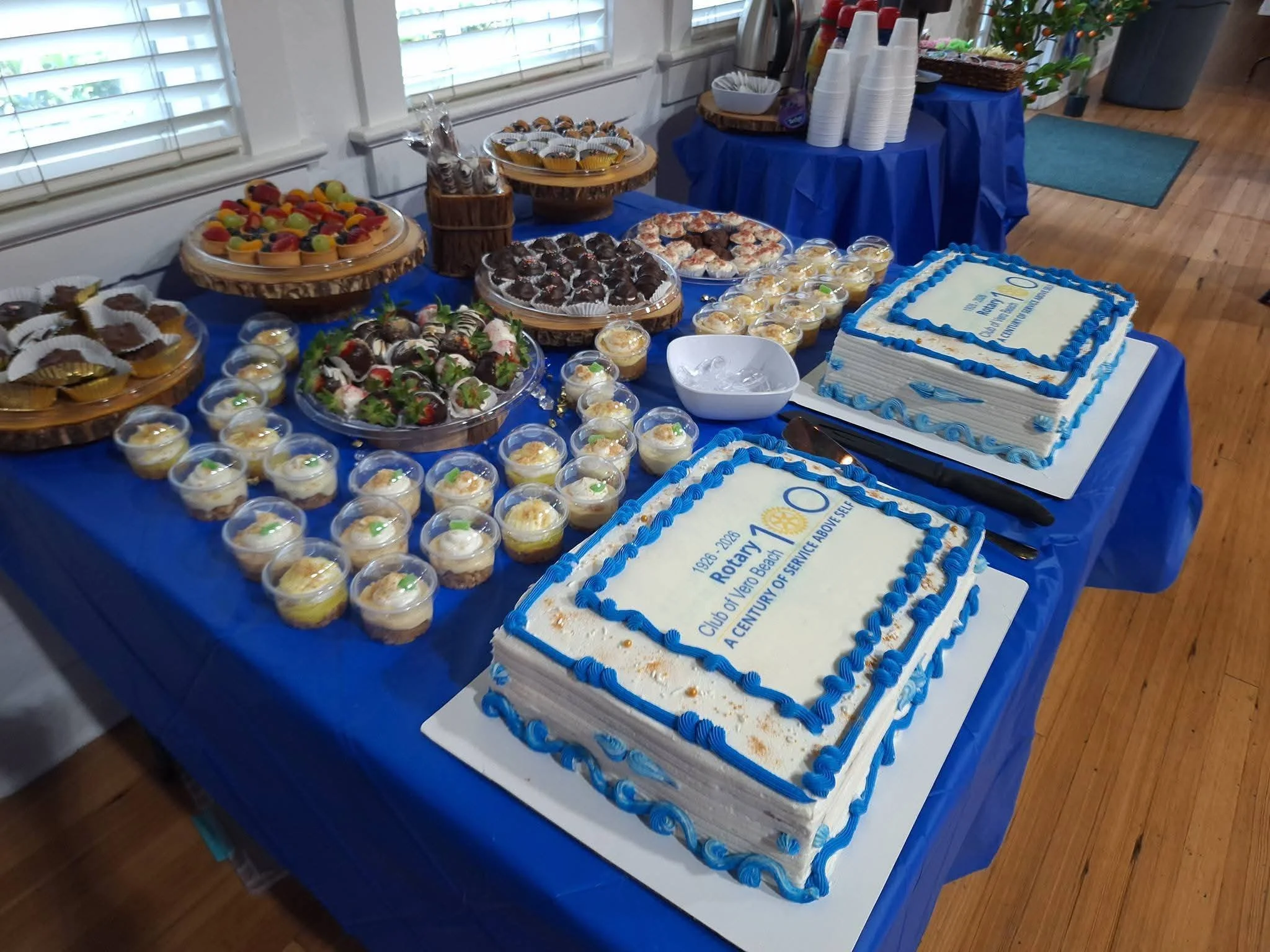 A table covered with a blue tablecloth displays an assortment of desserts including cupcakes, cookies, cake, and pudding, with a large rectangular decorated cake in the foreground celebrating a Rotary Club event.