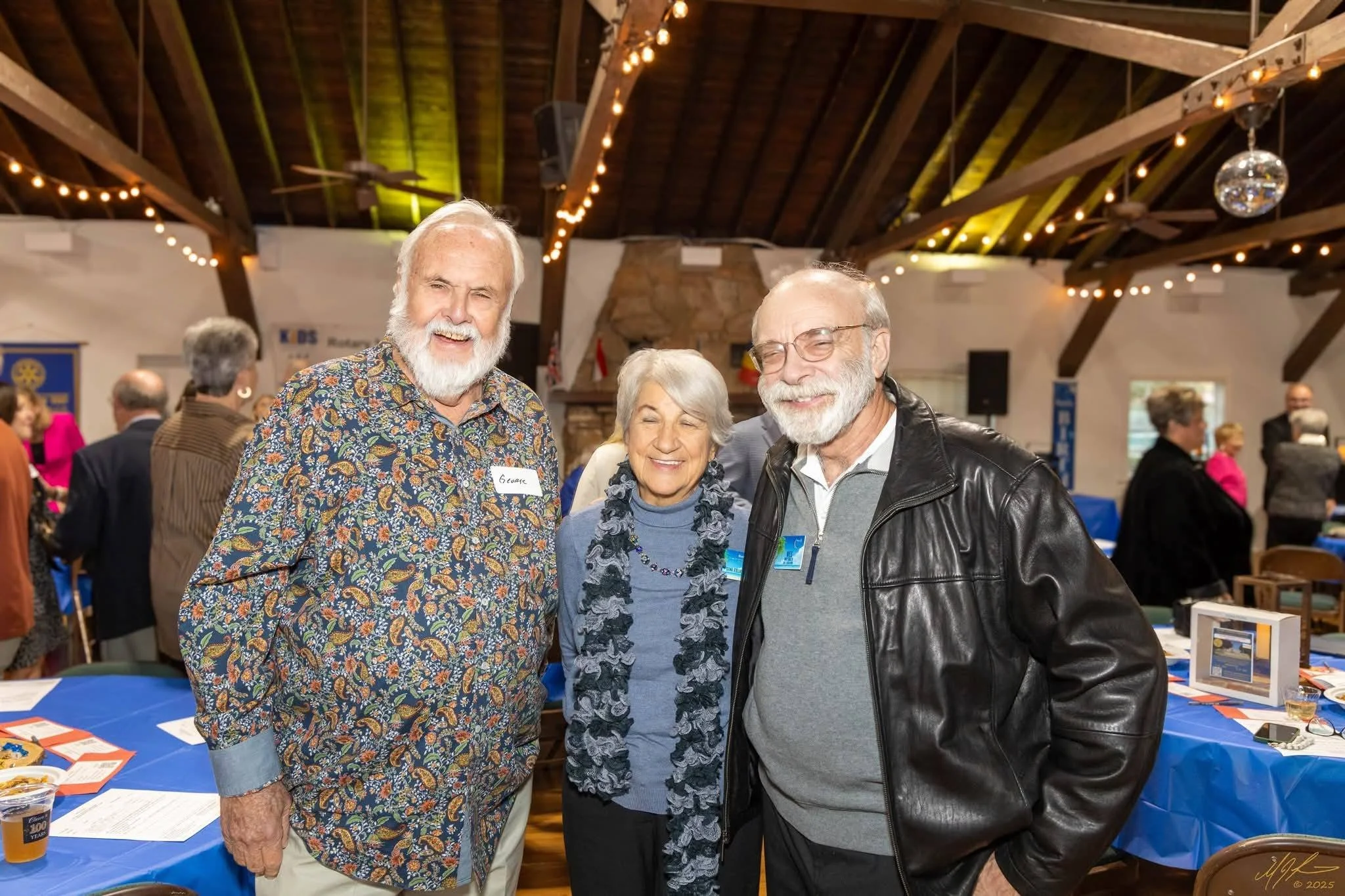 Four people smiling at an indoor event, three men and one woman, standing in front of a table with informational materials and decorations, with a rustic ceiling and string lights overhead.