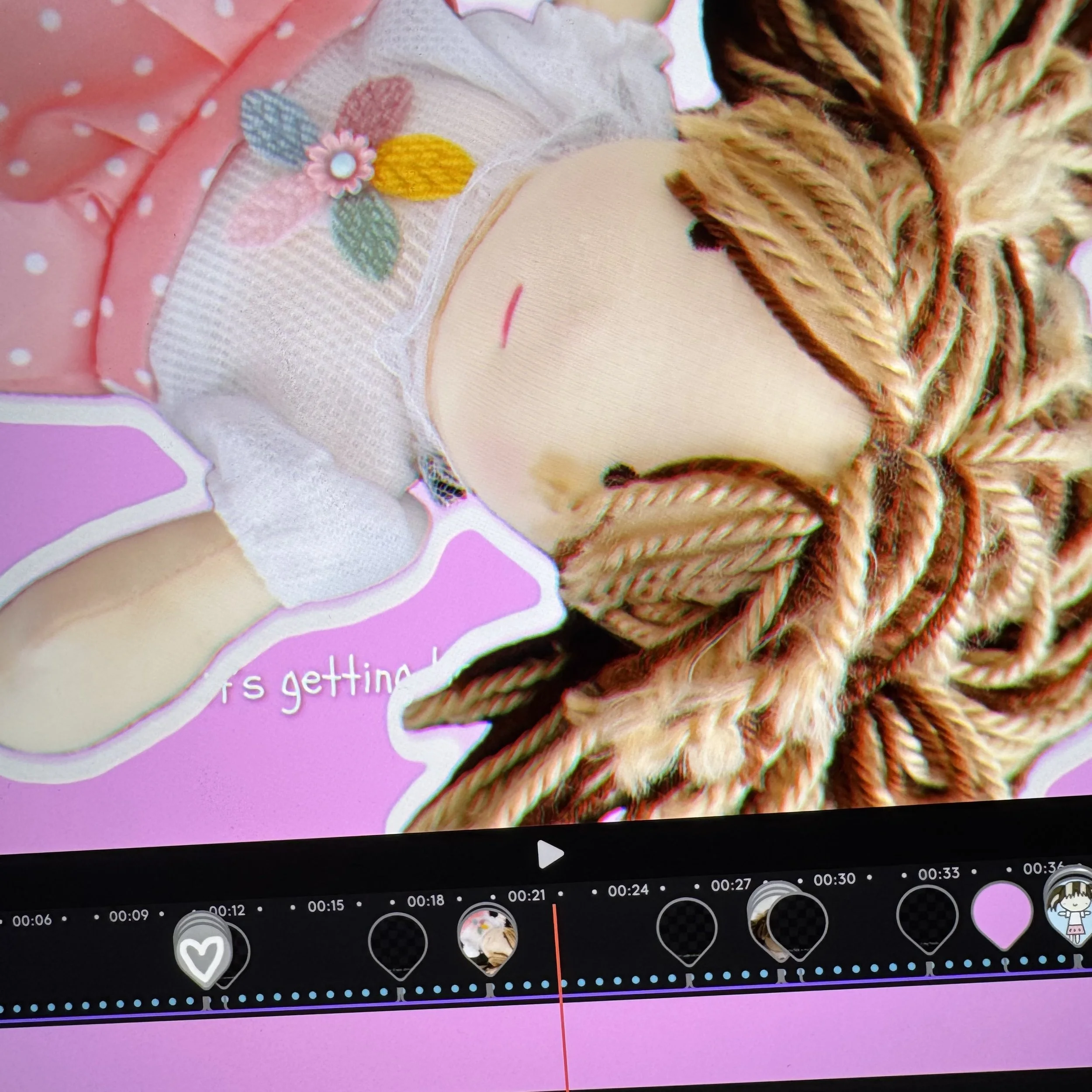 a doll with long braided hair, wearing a dress with floral embroidery, lying on a pink background showing a video editing timeline.