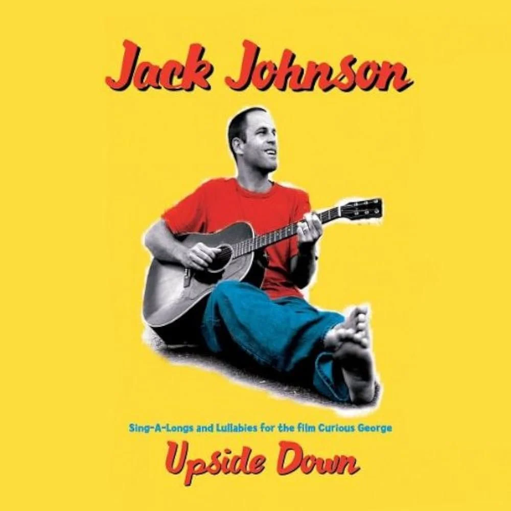 Album cover for Jack Johnson's 'Upside Down' featuring Jack Johnson sitting cross-legged, playing an acoustic guitar, wearing a red shirt and jeans, against a yellow background with red text.