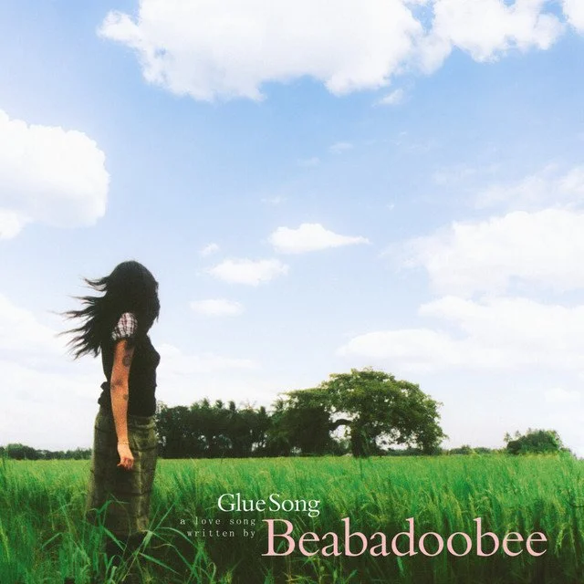 A woman with long dark hair standing in a green field under a blue sky with scattered clouds, facing sideways.