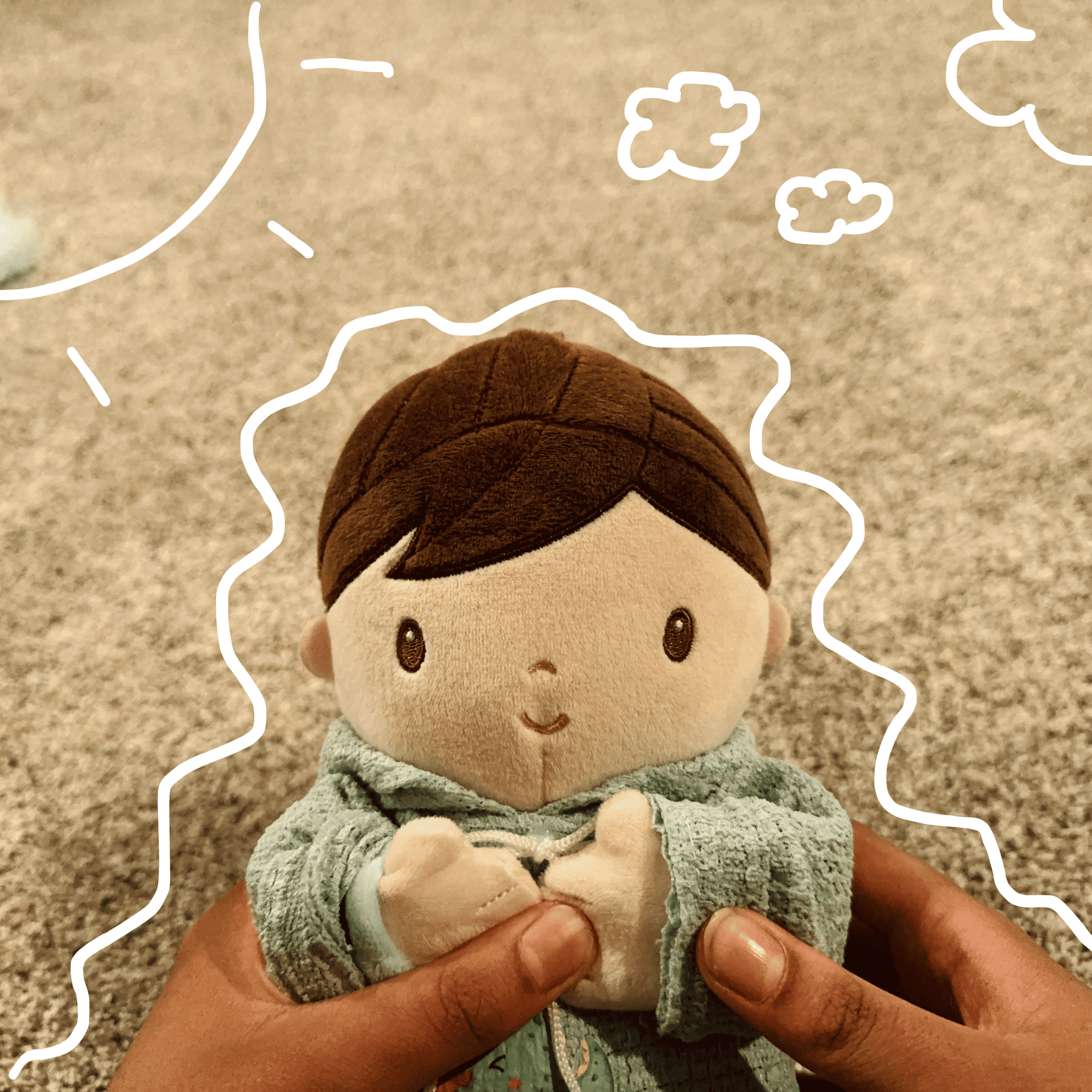 A plush doll with brown hair and a hoodie, held by two hands, on a textured beige surface with white chalk-like drawings of clouds and the sun around it.