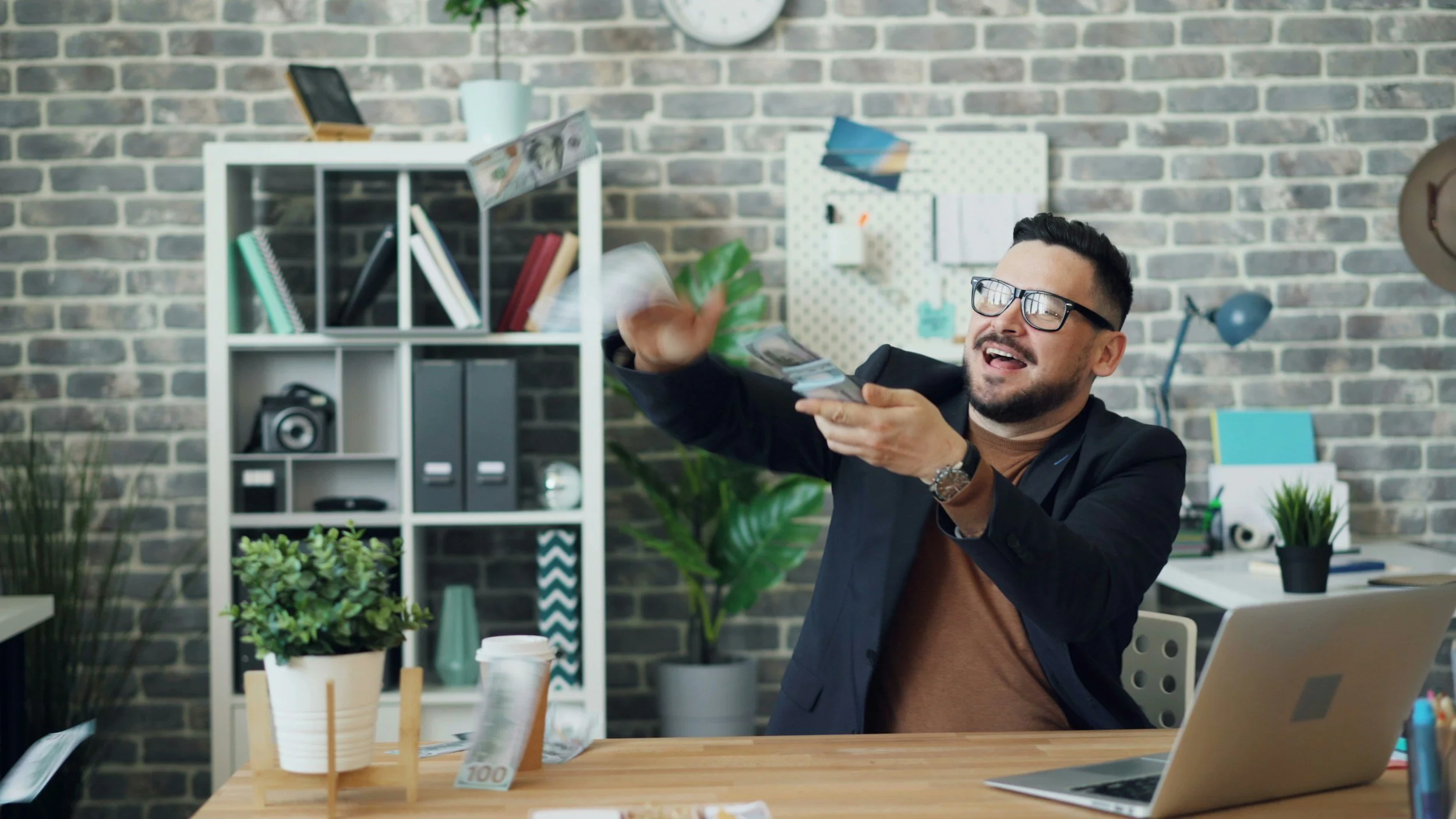 A man with glasses and a beard, dressed in a black blazer, sitting at a desk in a modern office with a brick wall background, excitedly tossing a stack of money into the air.
