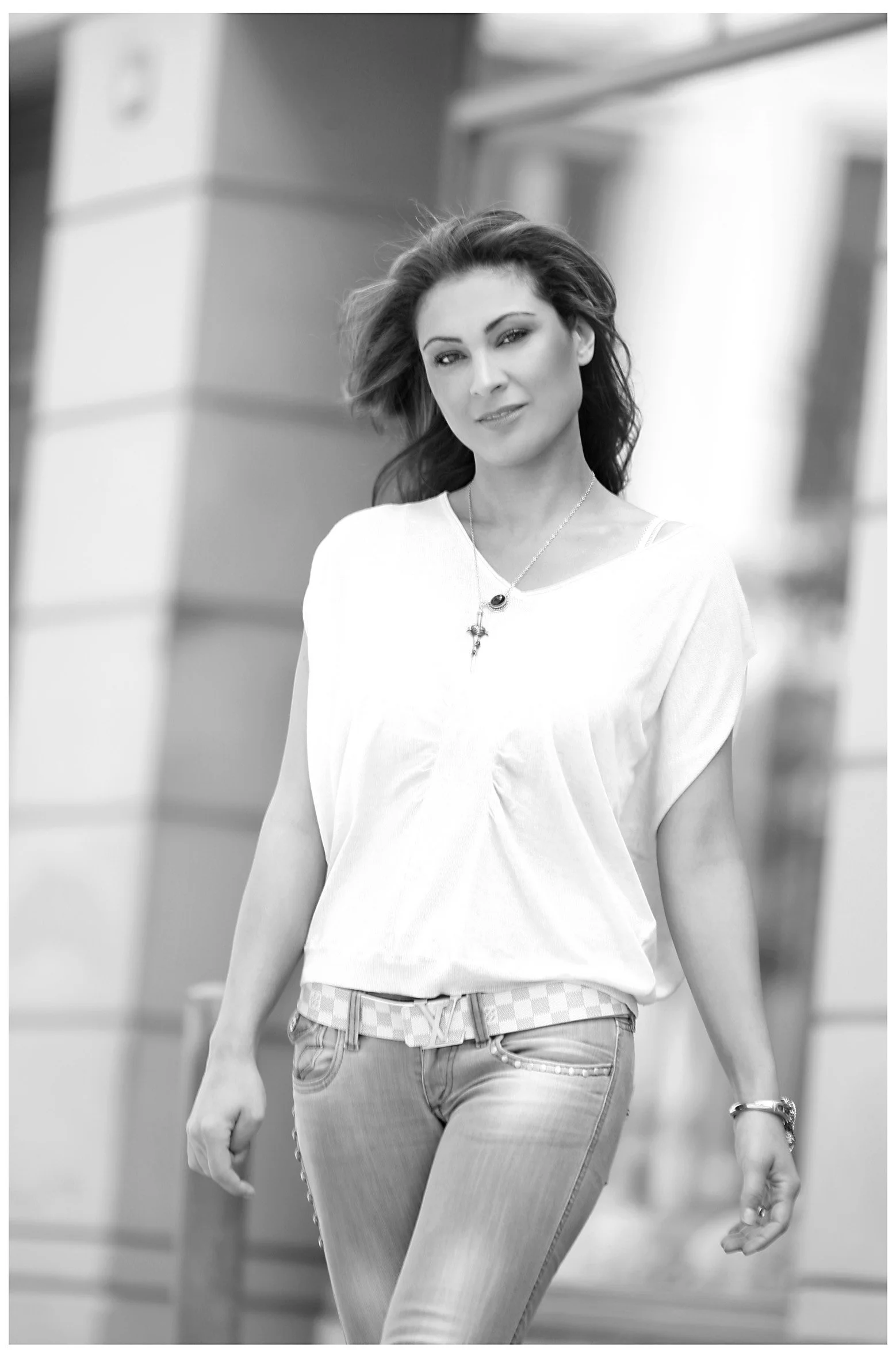 Black and white photo of a woman walking outdoors, wearing a light-colored top, jeans, and jewelry, with modern buildings in the background.