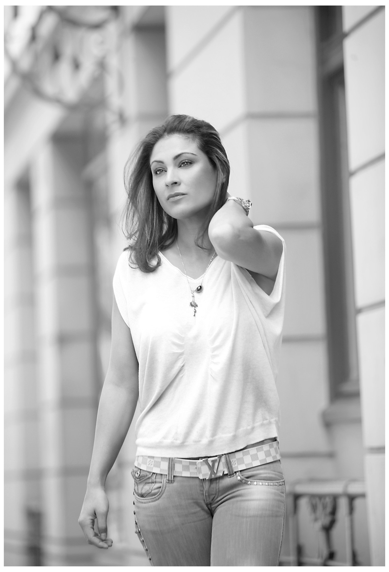 A woman with long hair, wearing a white top and jeans with a checkered belt, standing outdoors near a building with large windows, looking thoughtfully into the distance.