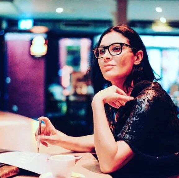 A woman with glasses sitting at a table in a restaurant or cafe, holding a pen and looking thoughtfully to the side.