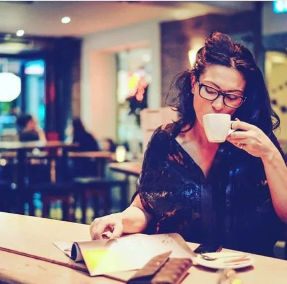 A woman with glasses and dark hair sitting at a table in a cafe, drinking from a white cup, with an open book or magazine on the table.