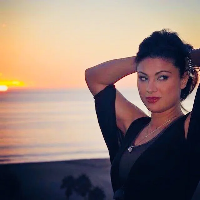 A woman with dark hair, wearing a black top, standing outdoors by the water during sunset, with a confident expression and her hand behind her head.