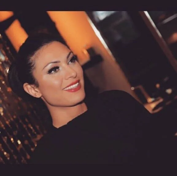 A woman with dark hair styled in an updo, wearing makeup with red lipstick, smiling, in a dimly lit indoor setting with warm lighting and decorative elements.