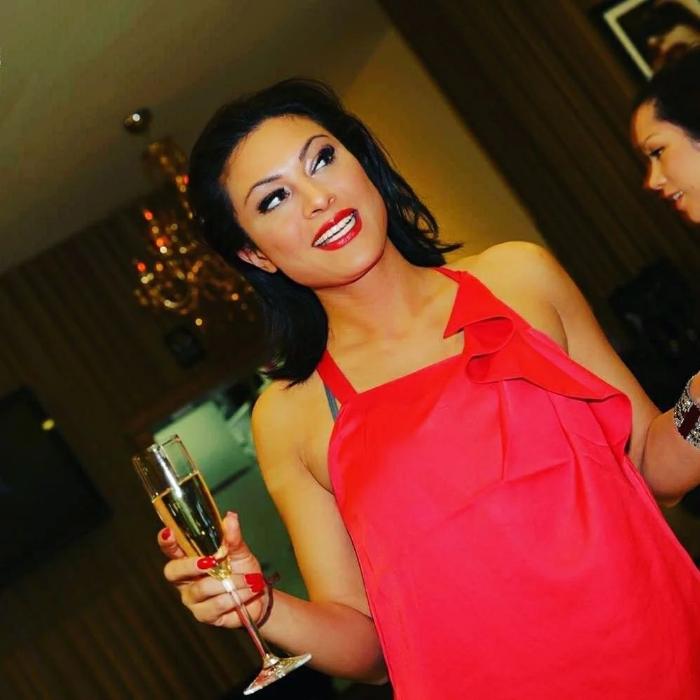 A woman with black hair and red lipstick wearing a red dress, holding a glass of champagne, at a social gathering.