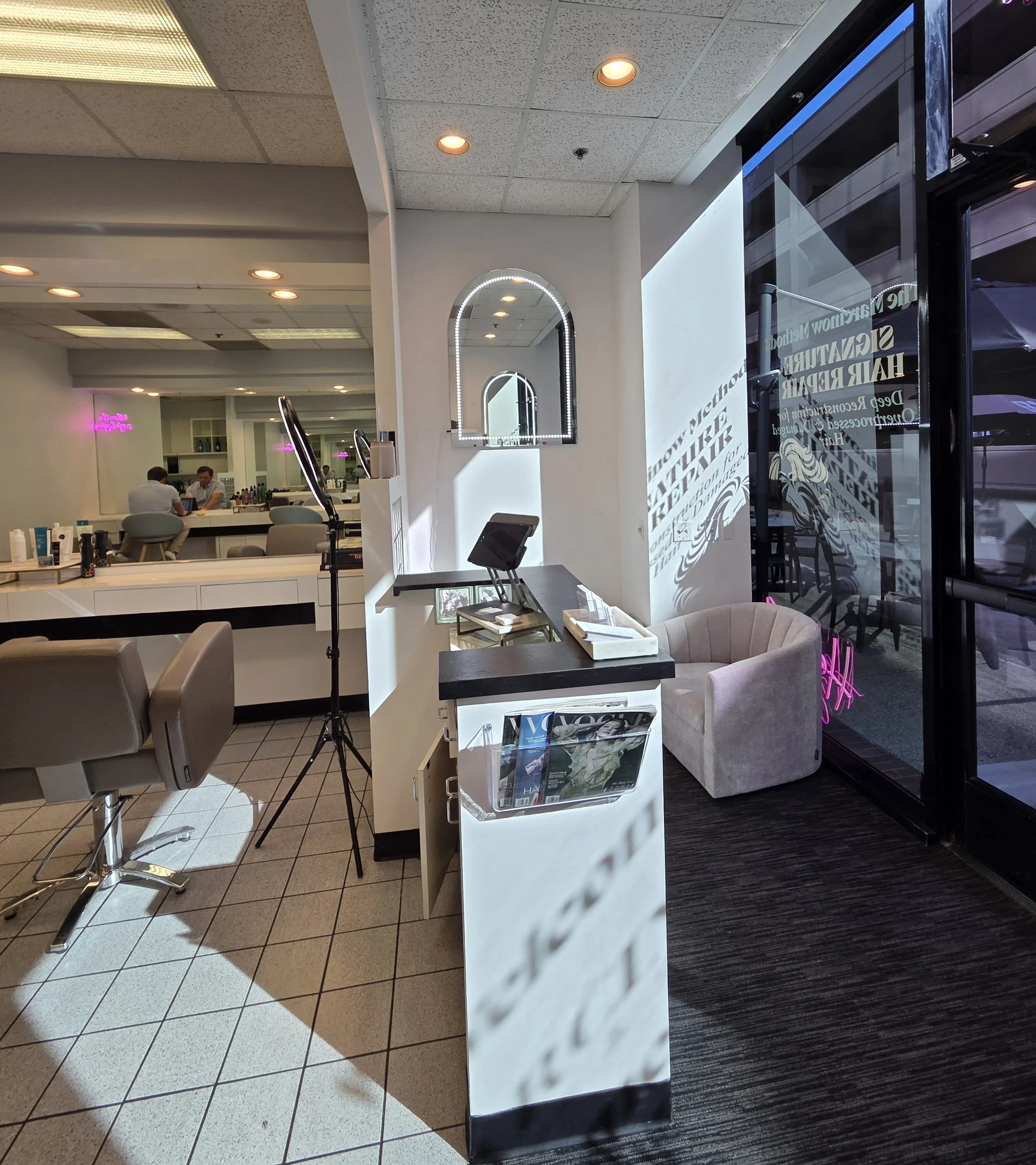 Interior of a modern salon or hair studio with chairs, a reception desk, a mirror on the wall, and a glass door with signage, sunlight streaming in.
