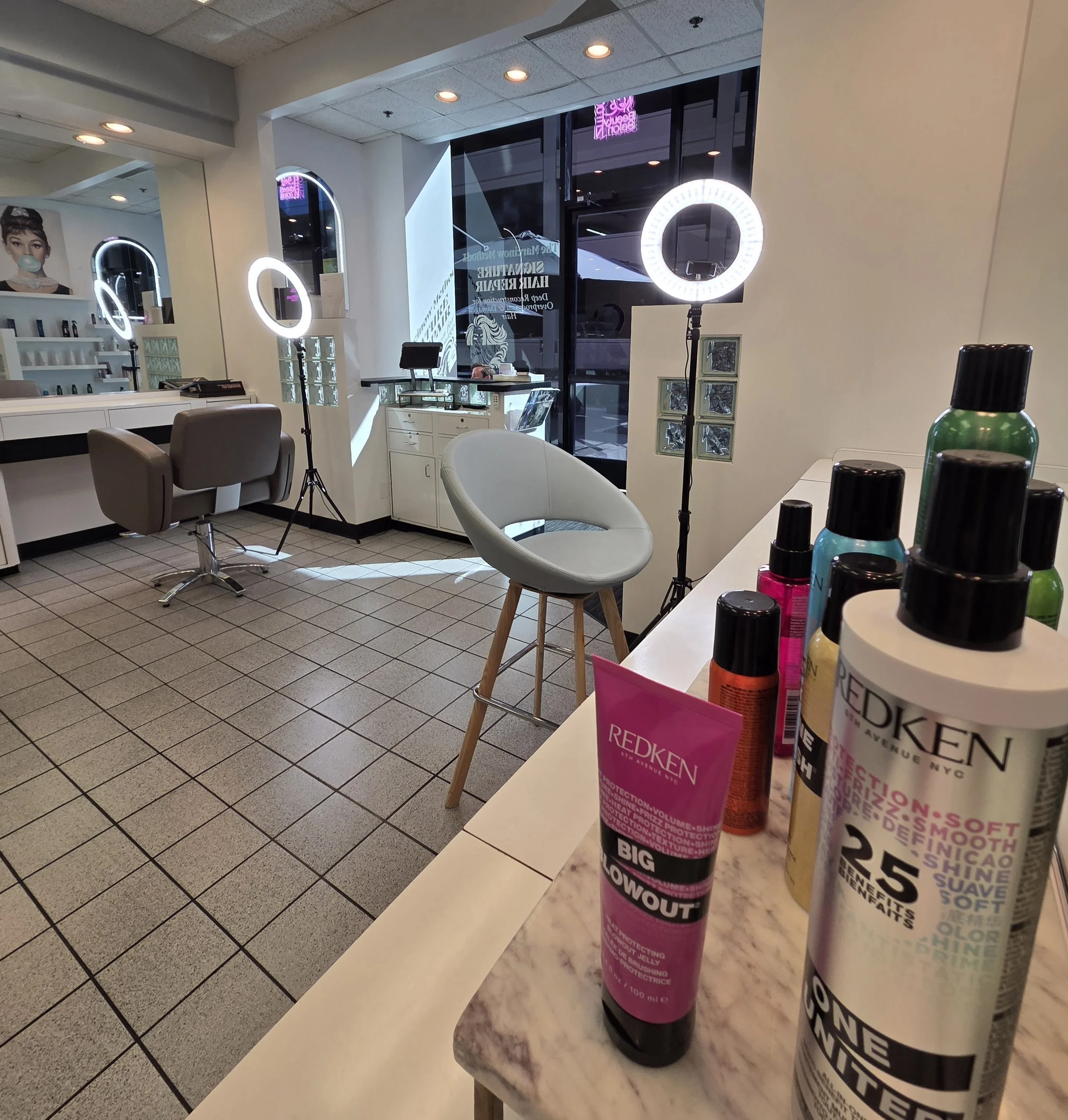 Interior of a beauty salon with salon chairs, ring lights for makeup or skincare, a mirror, and various hair and skincare products on a counter.