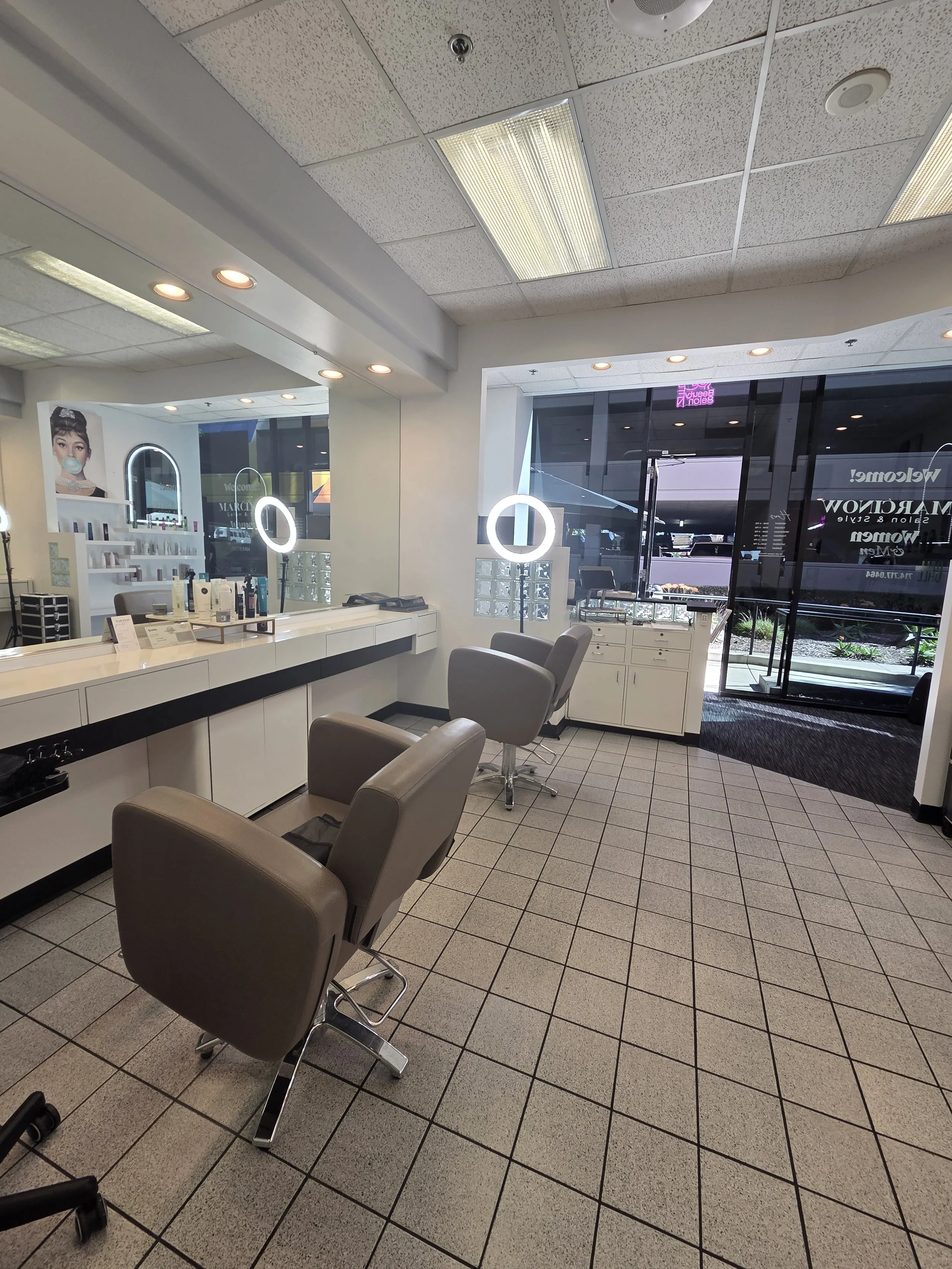Empty salon with three gray chairs, large mirror, bright lighting, glass door, and salon products on counter.