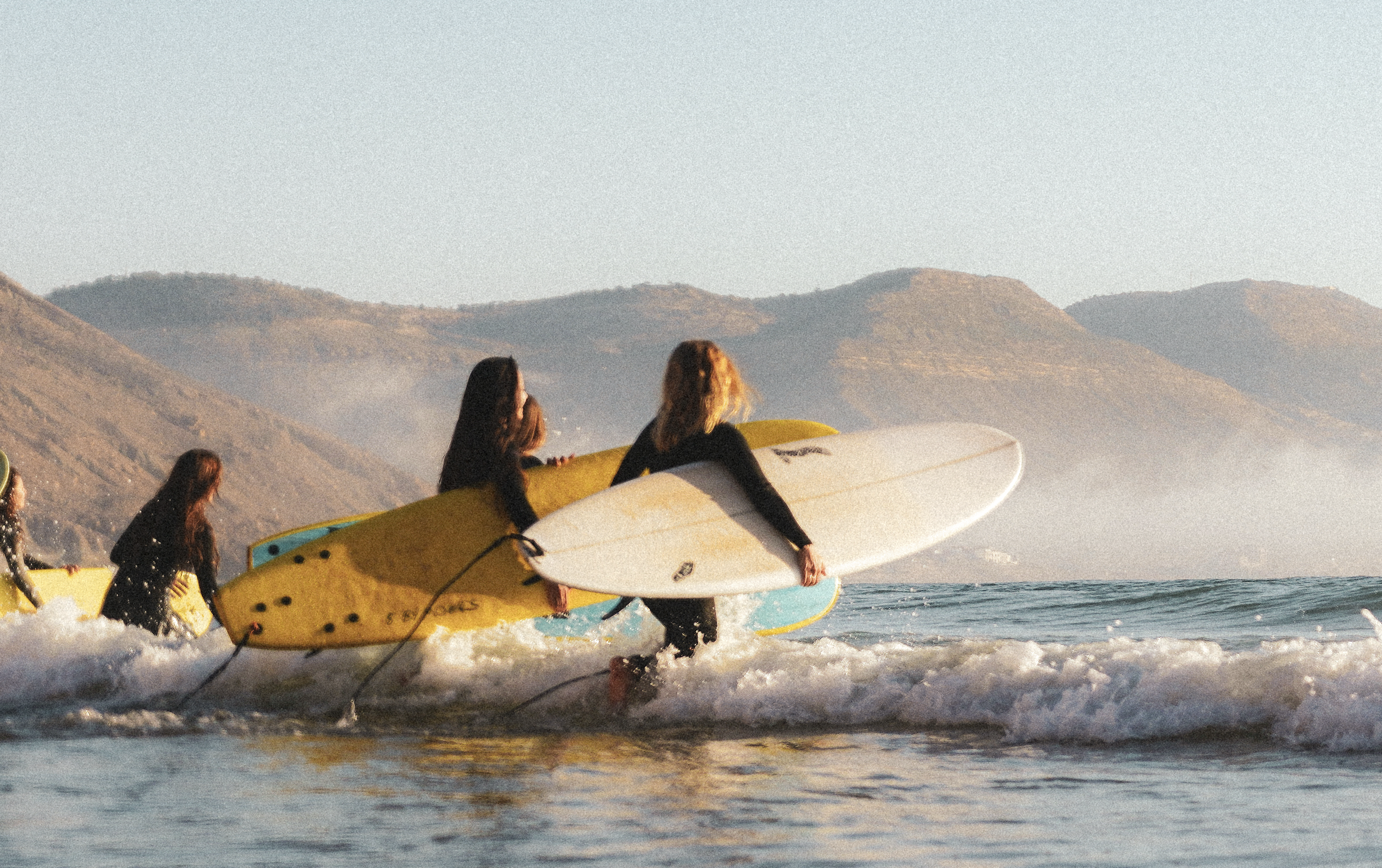 Surf lifestyle photography print of female surfers entering the ocean at sunrise with coastal mountains of imsouane Morocco
