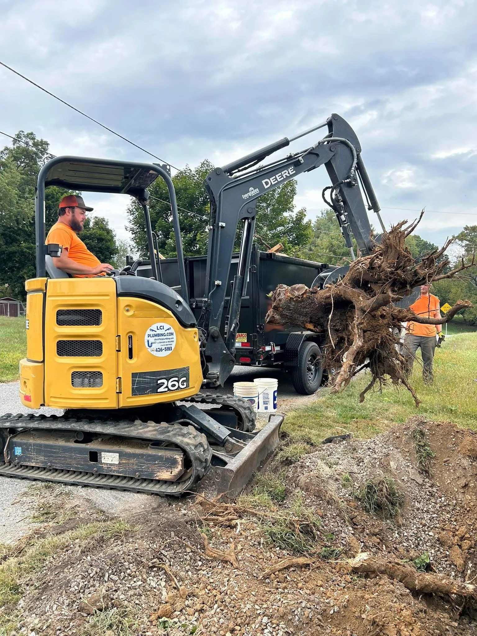 Person operating a small yellow excavator lifting a large uprooted tree trunk, with another person standing nearby on a grassy area.