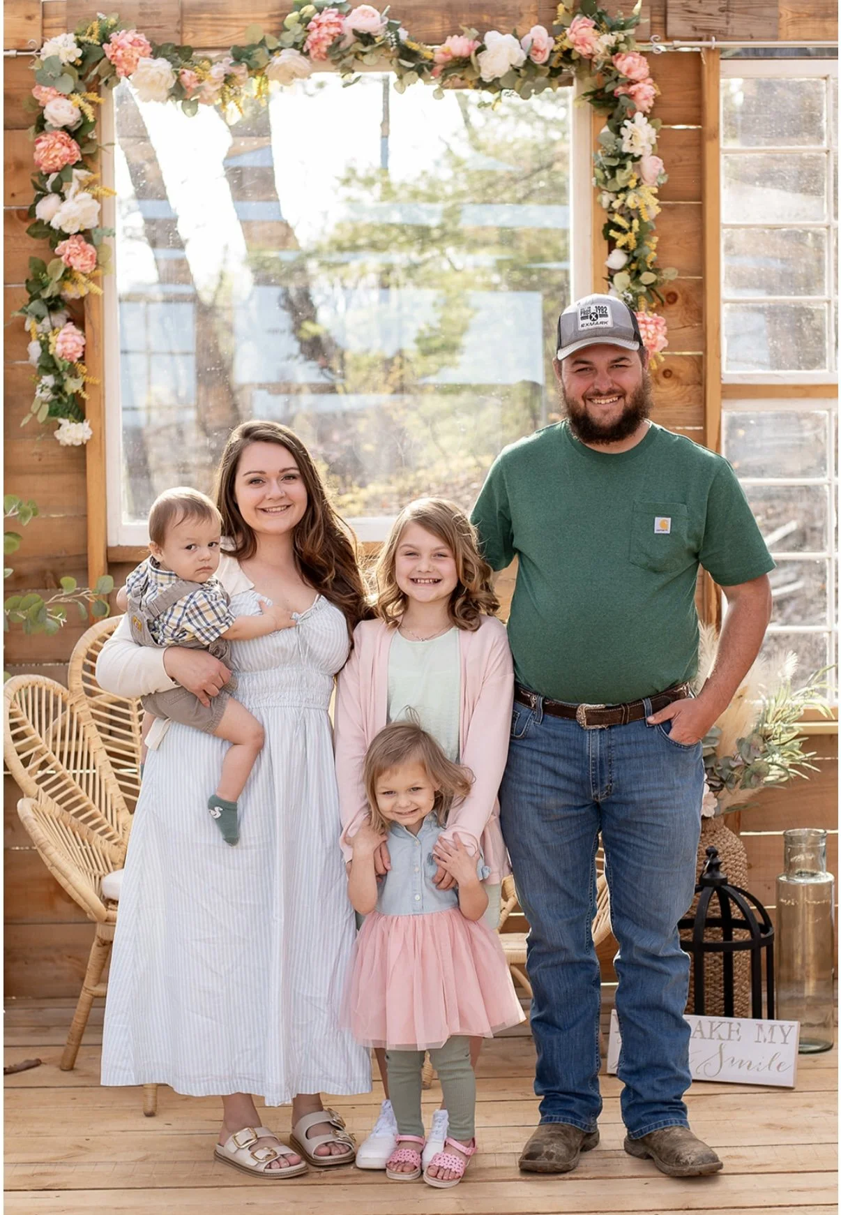 Family of five posing indoors with a wooden wall and large window decorated with a floral garland. The family includes a woman holding a young boy, a man, and three young girls. There is a sign on the floor that reads 'Take my smile.'