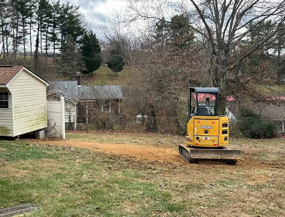 A small yellow Deere mini excavator parked on a dirt patch in a backyard with grass. There are houses, trees, and a cloudy sky in the background.