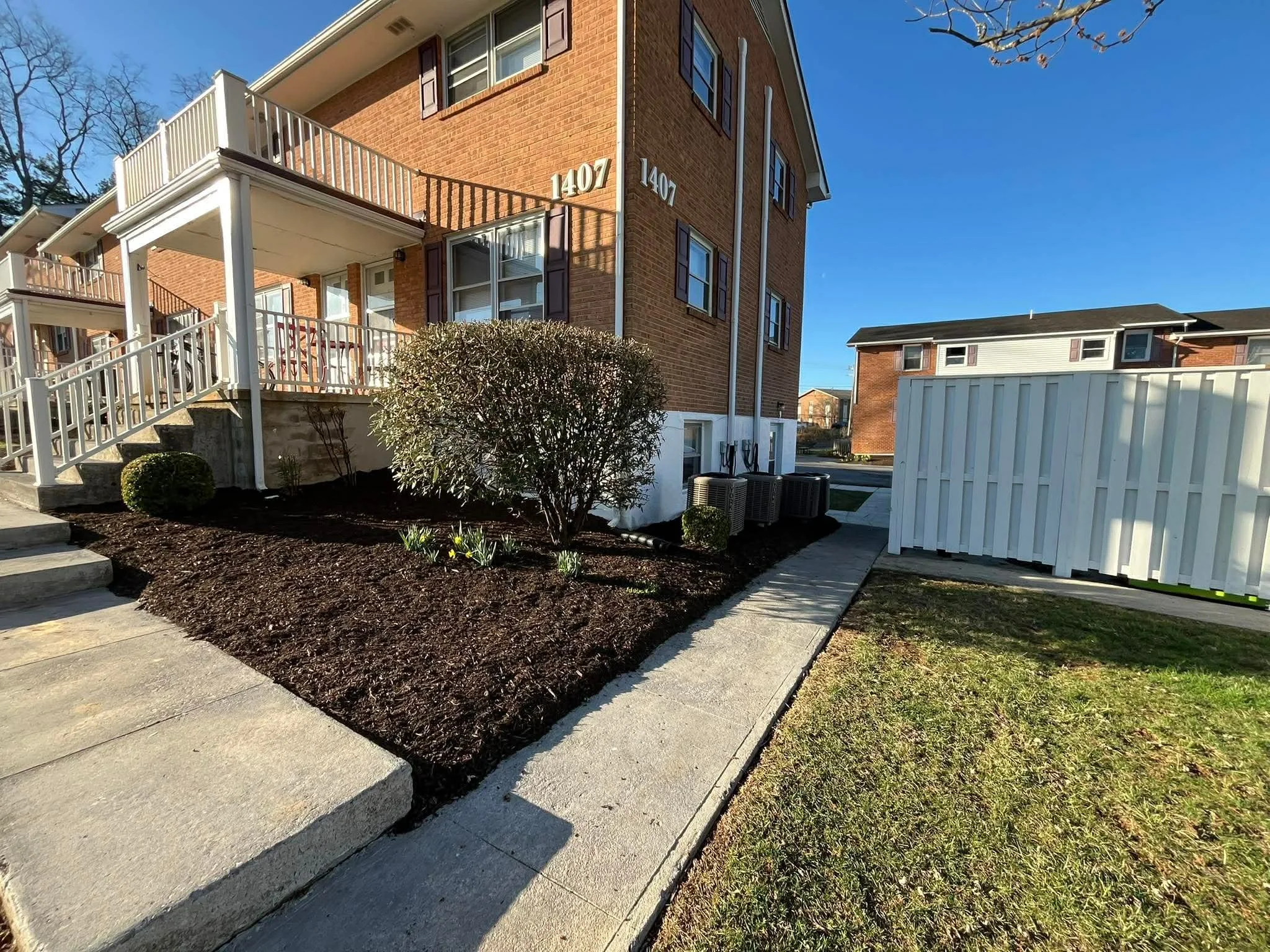 Exterior view of a brick apartment building with number 1407, showing a landscaped area with a bush, small plants, concrete sidewalk, and a white fence on a sunny day.