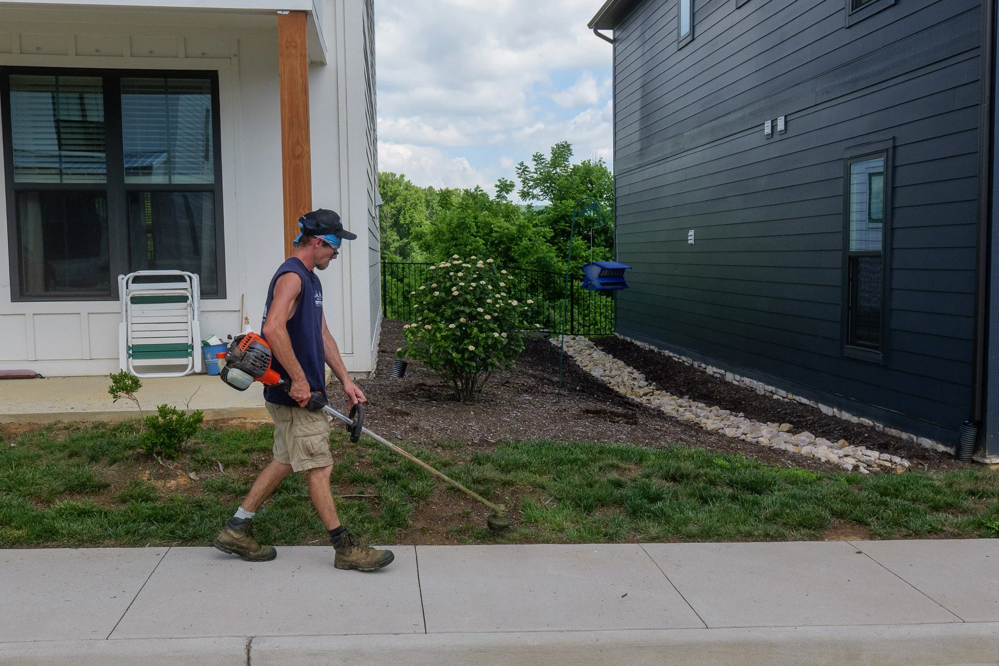 A man wearing a cap, sunglasses, sleeveless shirt, shorts, and boots is using a string trimmer to cut grass or weeds in a garden area between two houses. One house has white siding and the other has dark gray siding. There are bushes, a flower, and landscaping rocks, with trees and a cloudy sky in the background.