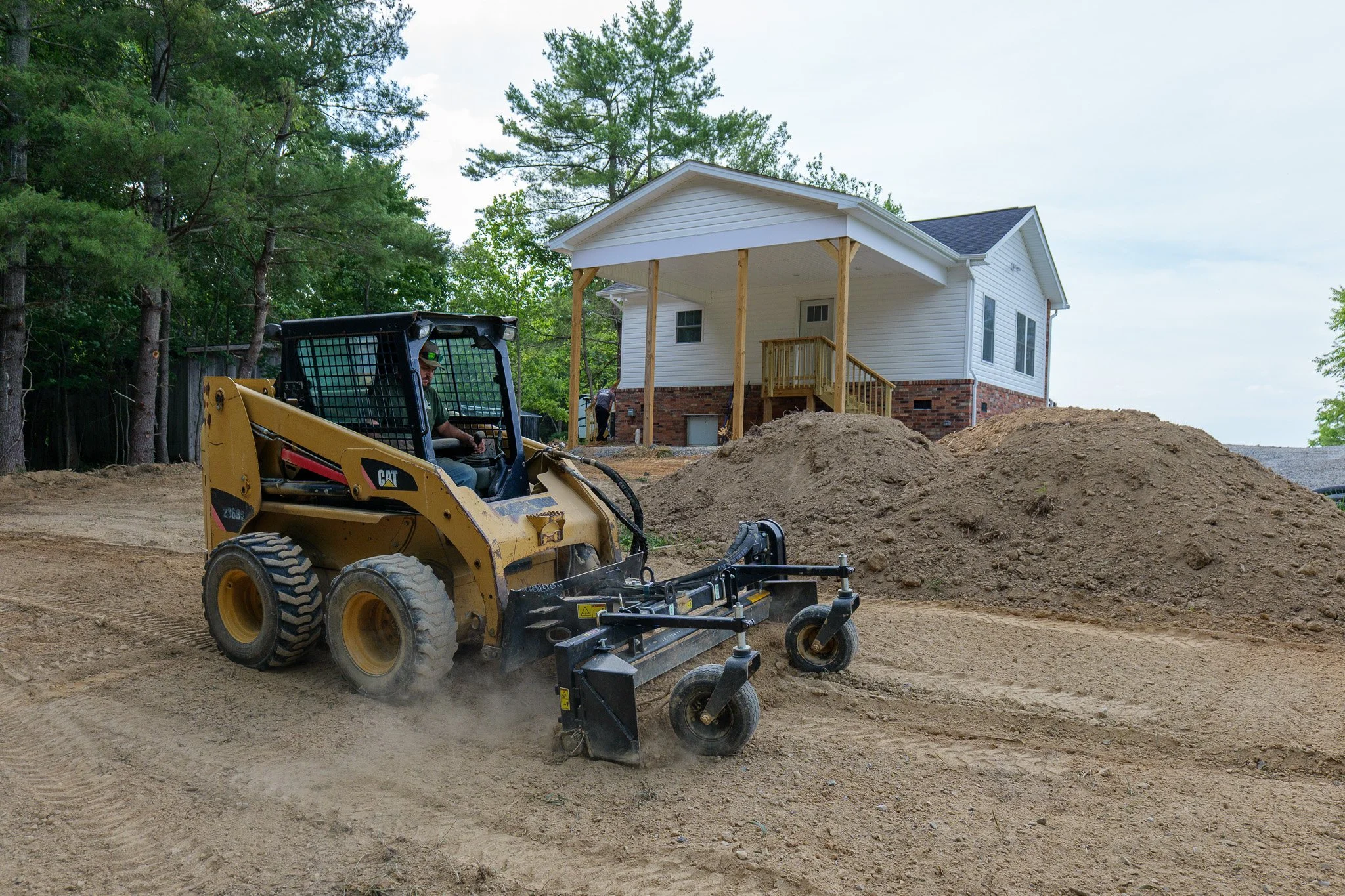 Construction worker operating a yellow compact tractor on a dirt construction site, with a white house with a small porch and a brick foundation in the background, surrounded by trees.