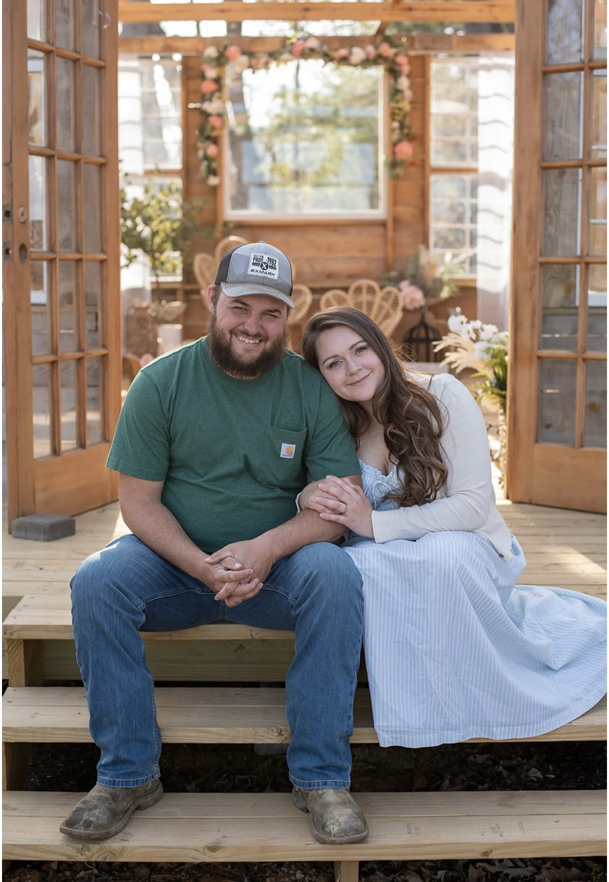 A man and woman sitting on wooden steps, smiling at the camera inside a greenhouse or garden room decorated with flowers and wooden furniture.