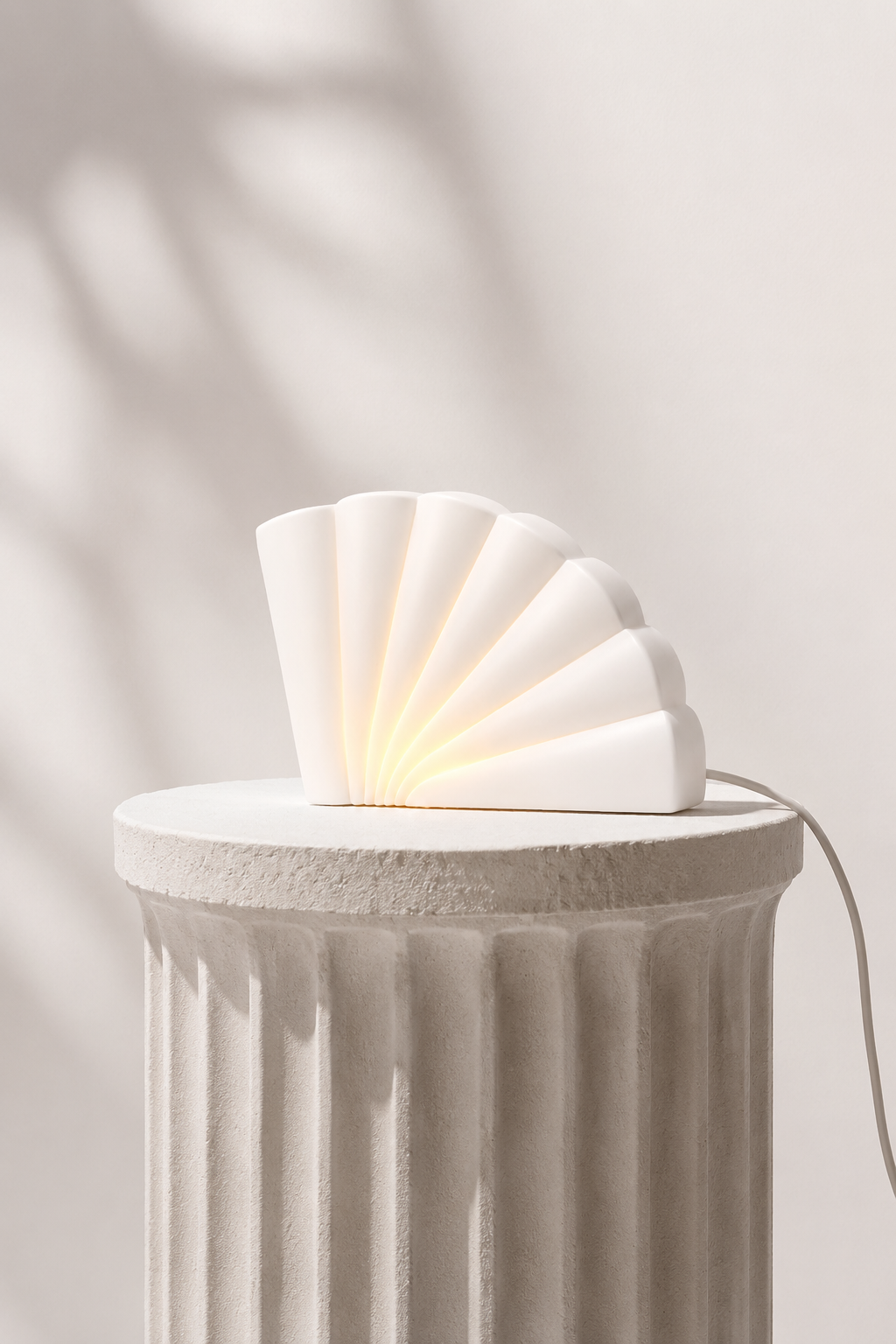 A modern, white, shell-shaped lamp with glowing warm light, placed on a textured, off-white pedestal against a light background with soft shadows.
