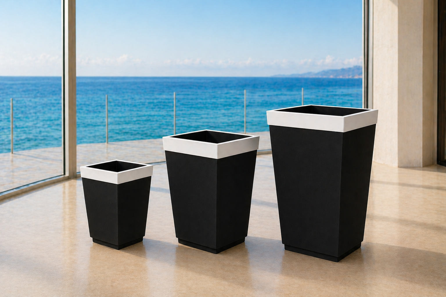 Three large black and white empty flower pots of varying sizes on a beige floor near large glass windows overlooking a blue ocean and sky.