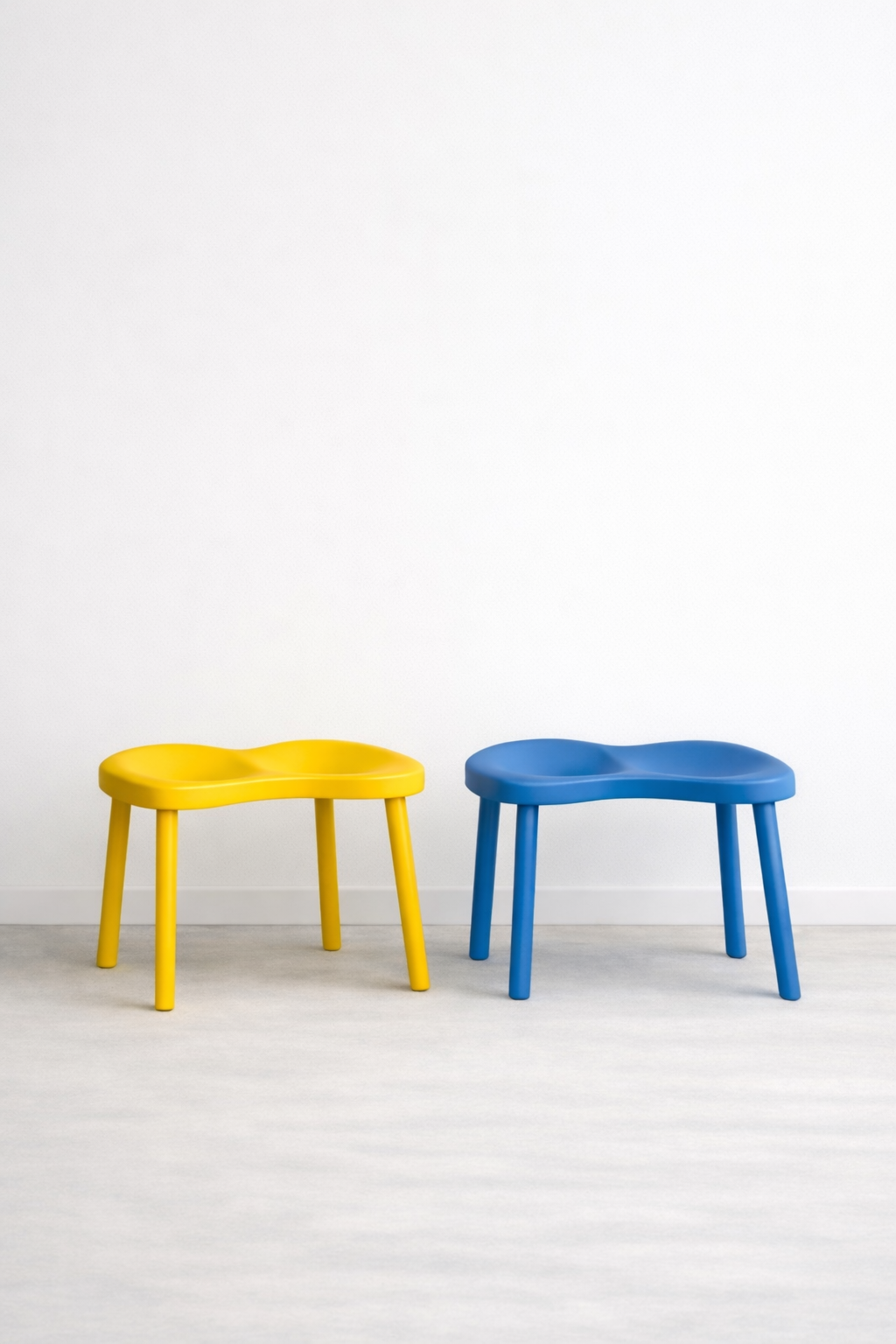 Two colorful children's stools, one yellow and one blue, placed against a plain white wall on a light-colored floor.