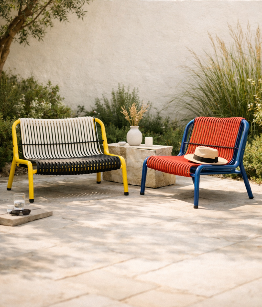Two colorful outdoor chairs, one yellow and black and the other blue and red, are positioned on a stone patio with a beige wall and greenery in the background. A small stone table between them holds a white vase with dried flowers, a small white cand