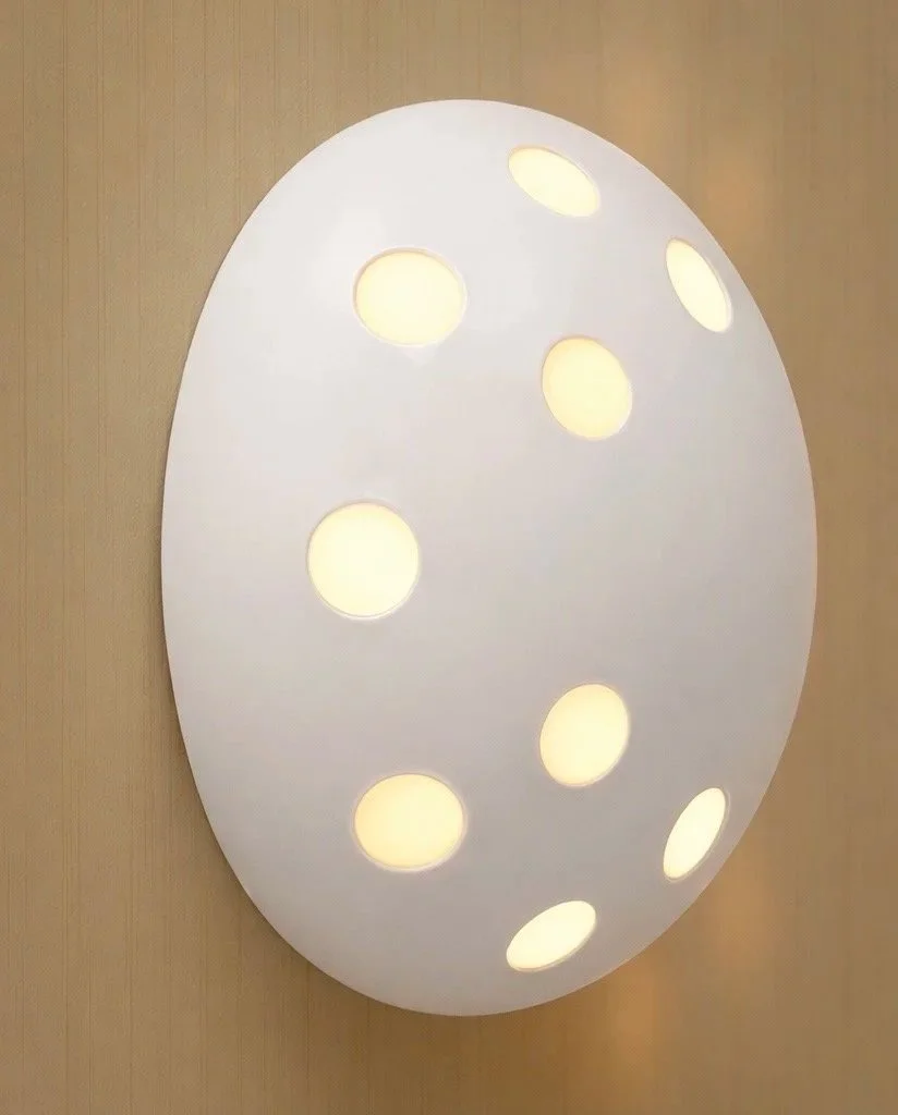 White circular wall light with multiple round cutouts emitting warm light, mounted on a beige wall.