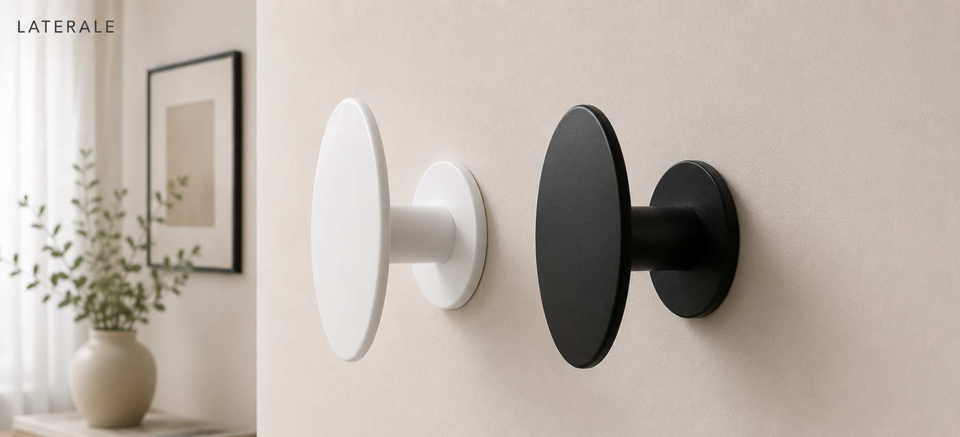 Two modern wall hooks, one white and one black, mounted on a beige wall, with a framed picture and potted plant in the background.