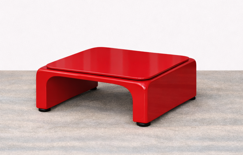 A modern red coffee table with curved edges on a grey carpeted floor against a plain white wall.