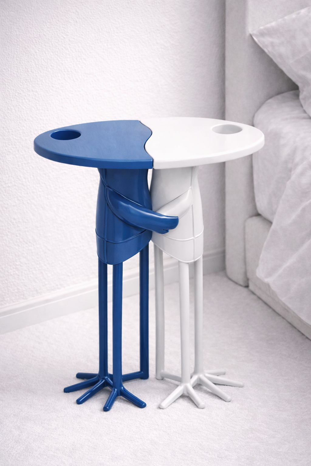 Two abstract human-shaped side tables, one blue and one white, with round tops and cup holders, placed beside a bed.