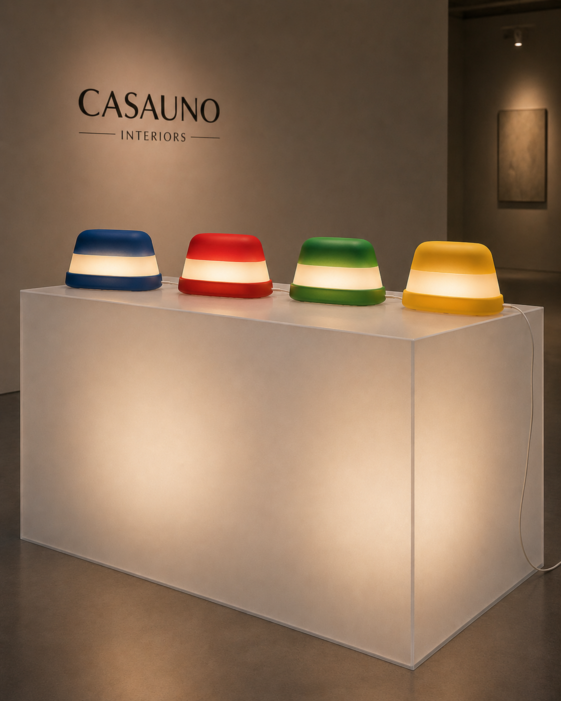 Four colorful table lamps in blue, red, green, and yellow are displayed on a white pedestal in an art gallery, with a wall sign reading 'CASAUNO INTERIORS' in the background.